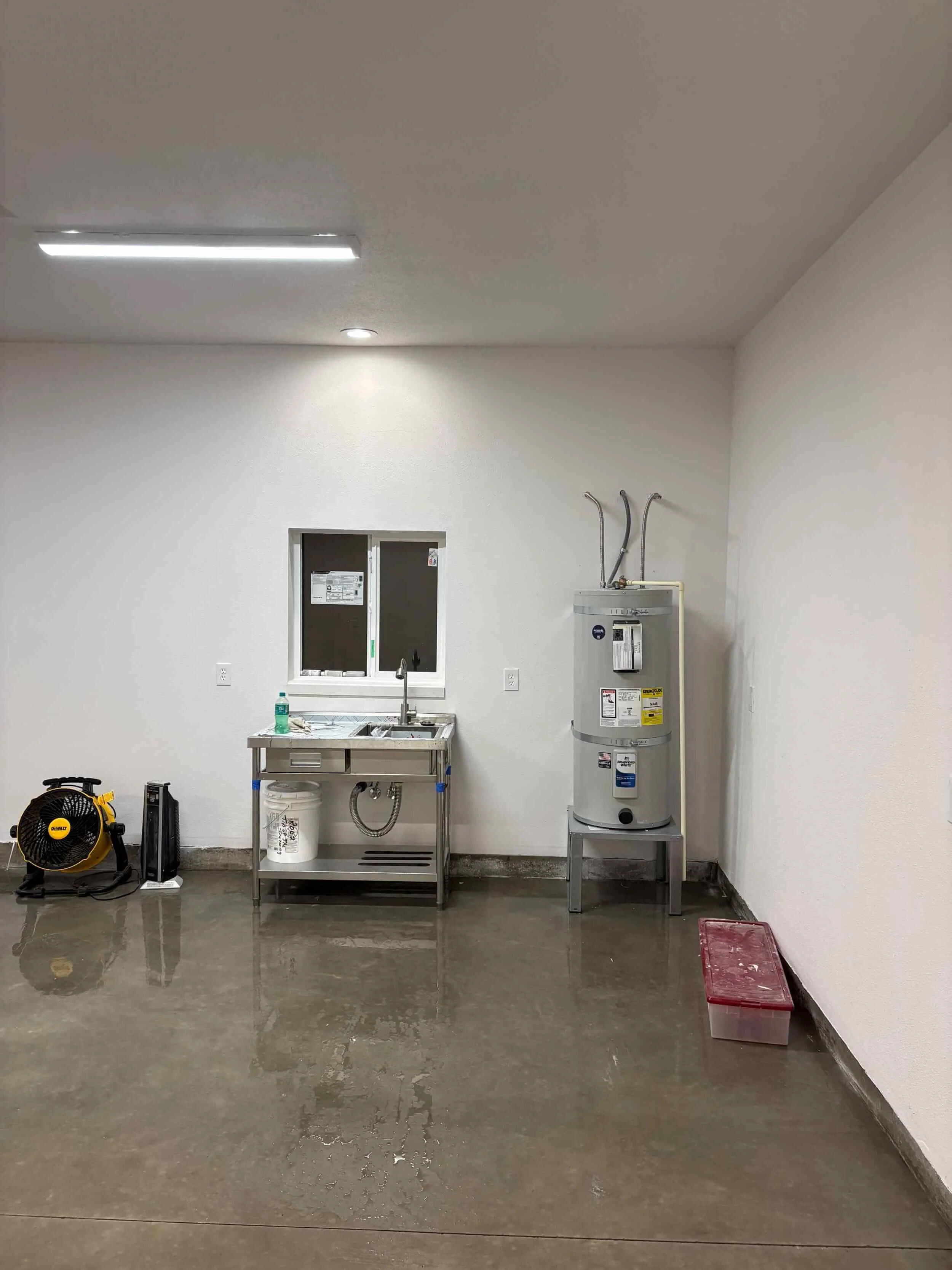 Empty utility or maintenance room with a concrete floor, white walls, and ceiling. Contains a black and yellow portable fan or air compressor, a small stainless steel sink, a water heater, and a red plastic storage bin.