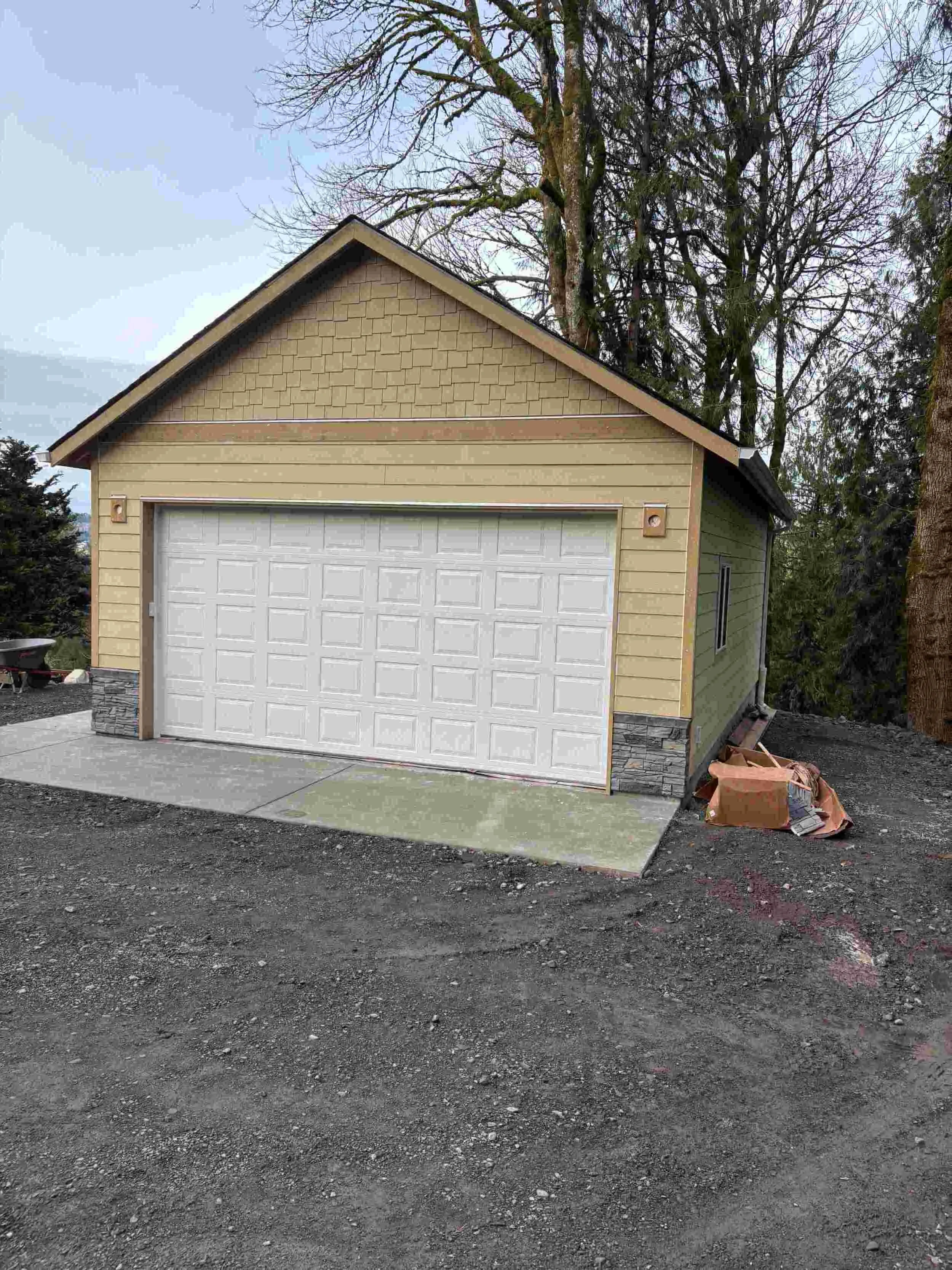 A small detached garage with beige siding and stone accents at the base, white garage door, and a concrete driveway in front. There are tall trees in the background and some construction debris on the side.