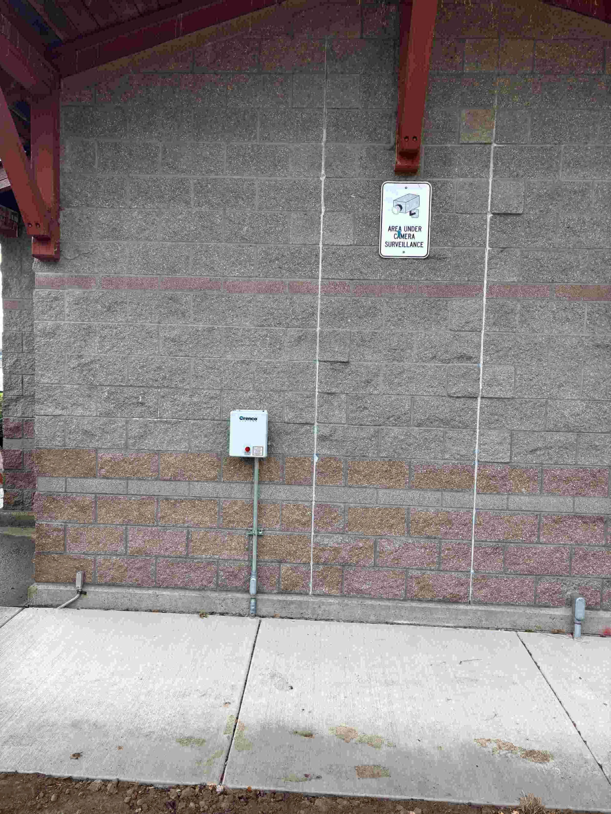 A gray brick wall with a surveillance camera sign, a white electrical box, and a small pipe near the ground. The camera surveillance sign reads 'AREA UNDER CAMERA SURVEILLANCE.'
