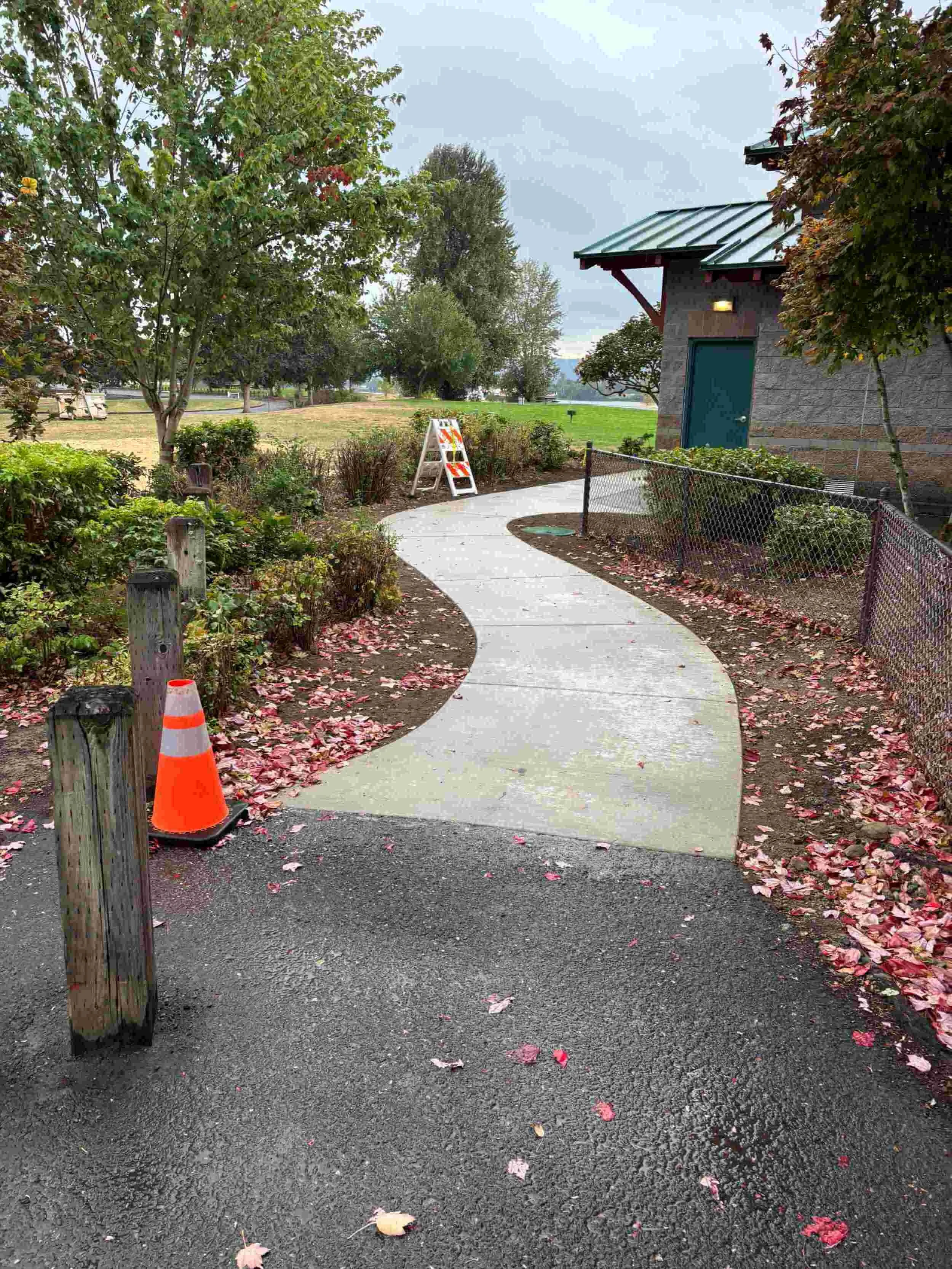 A winding concrete pathway through a landscaped park area with trees, bushes, fallen pink leaves, a chain-link fence, and a building with a blue door and green roof in the background. Orange traffic cone and signs are near the pathway.