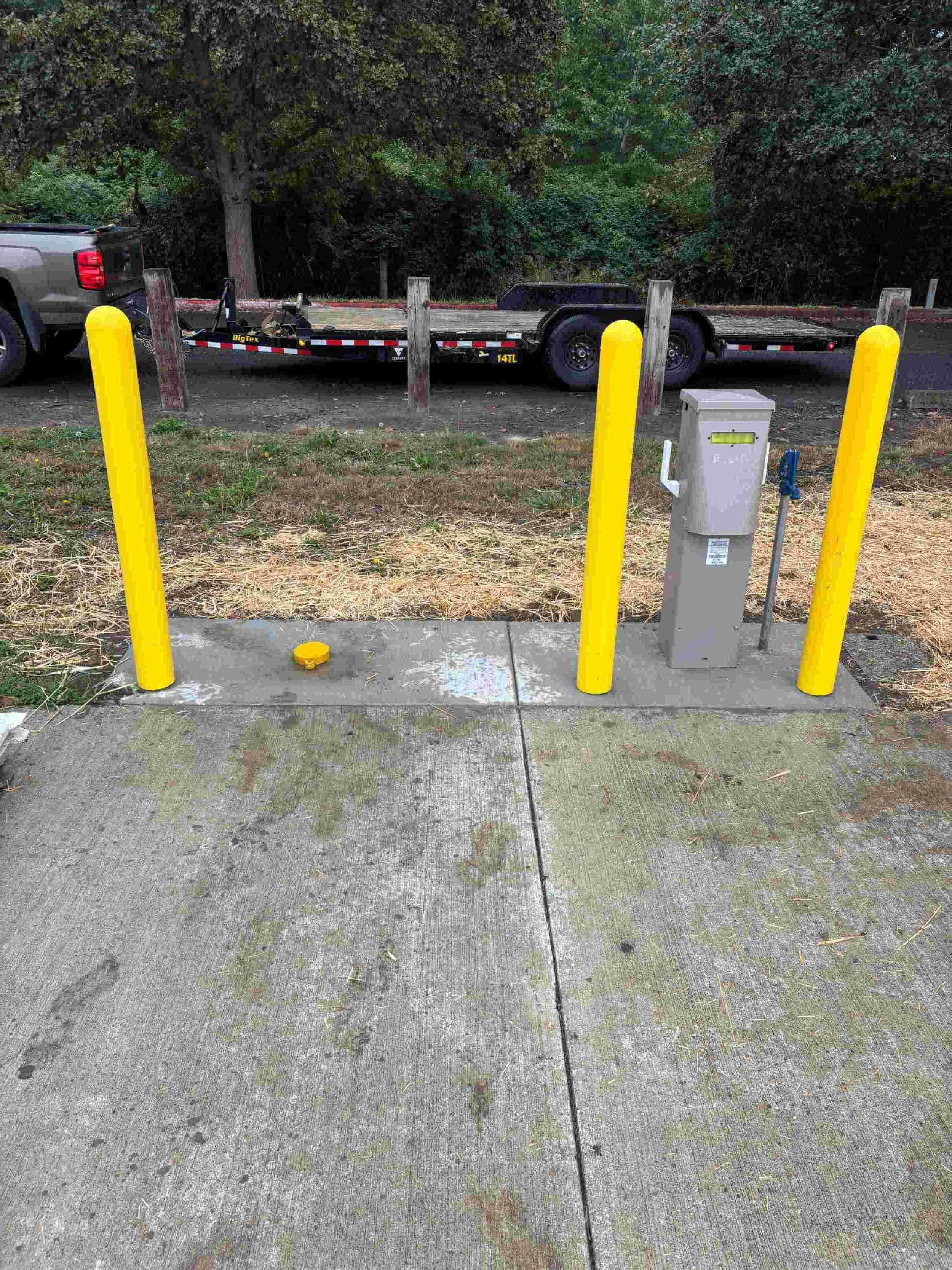 Sidewalk with yellow protective posts, electrical box, and metal pole, with a trailer and pickup truck in the background.