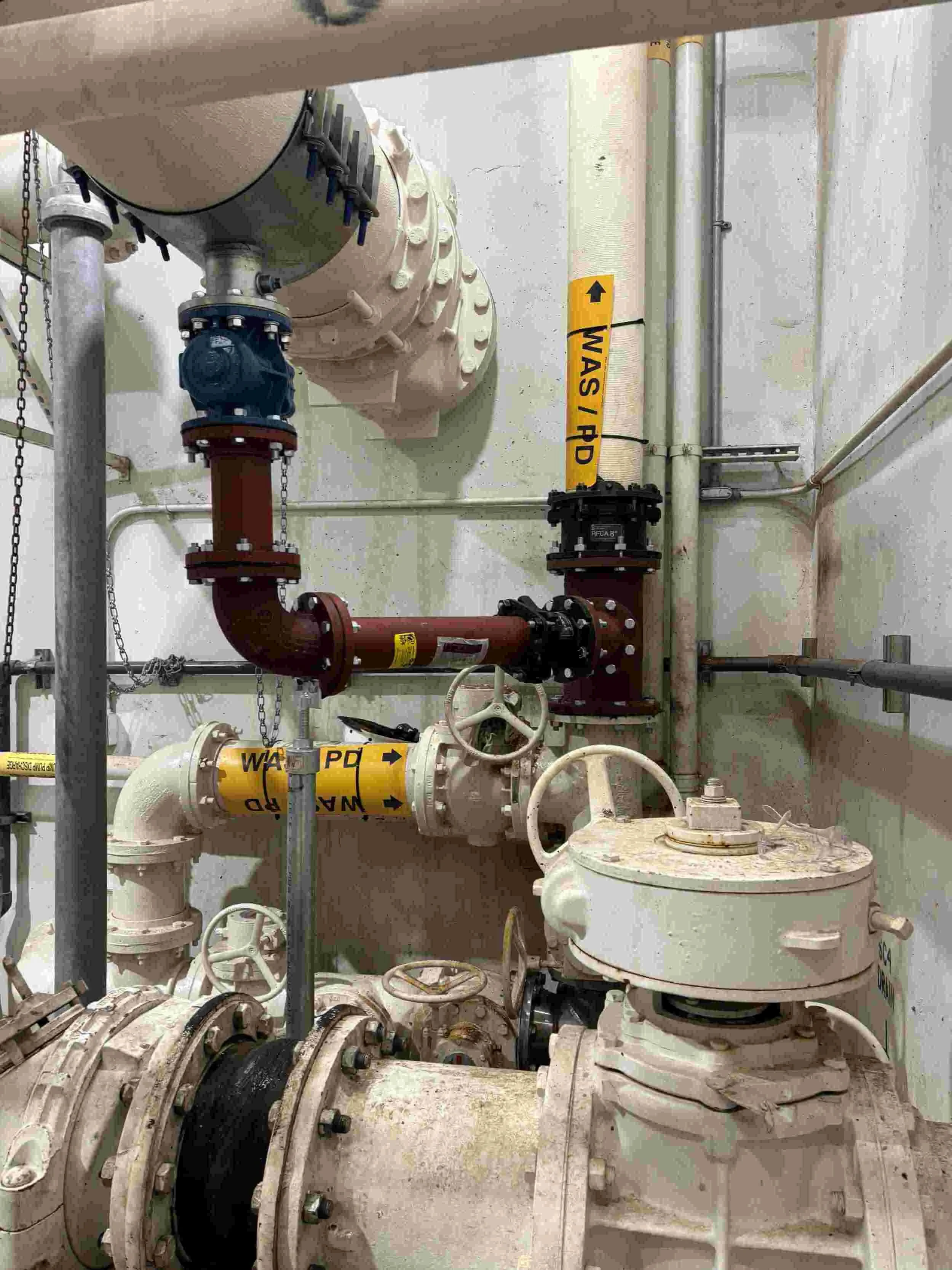 Industrial pipes and valves in a utility room, with labels such as WAS/PD on the pipes.