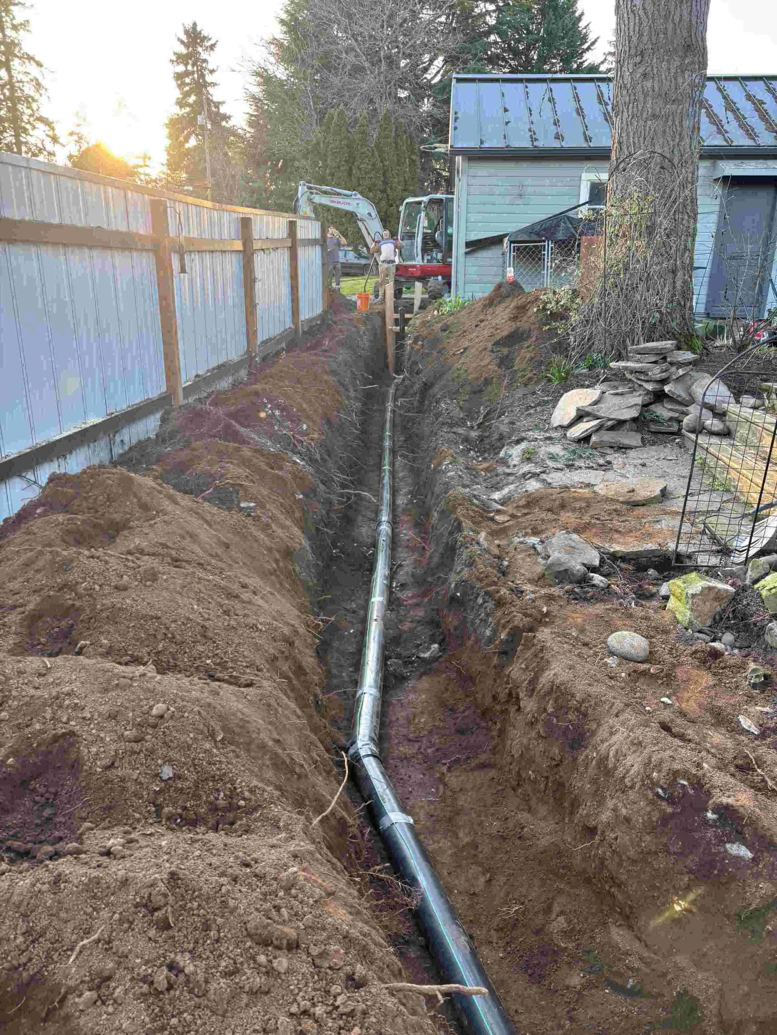 Workers installing or repairing a pipe in a trench in a backyard, with a house and trees in the background, during sunset.