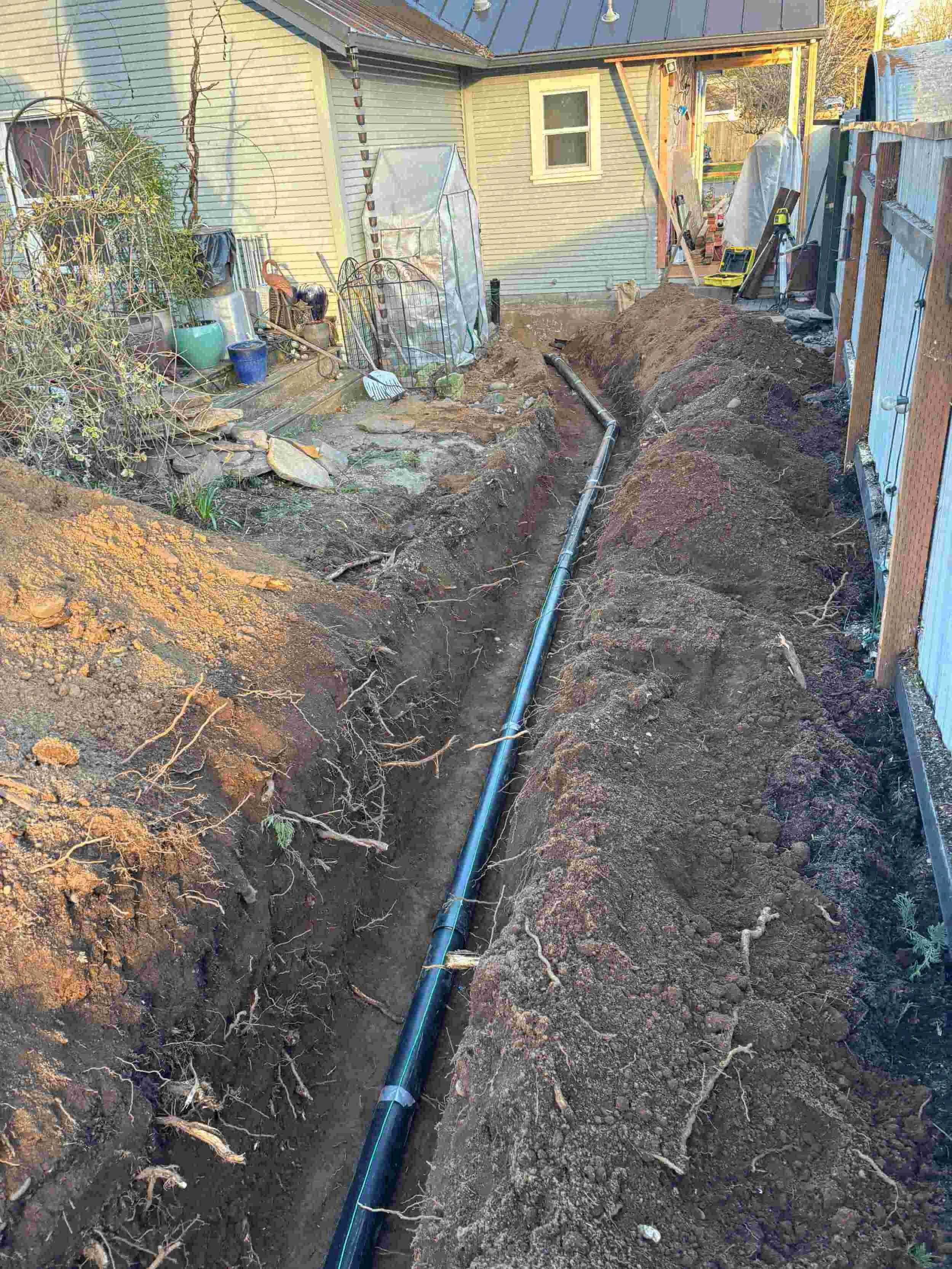 A backyard with an ongoing underground pipe installation, trenches dug in the ground for piping, a house, and construction tools and materials nearby.