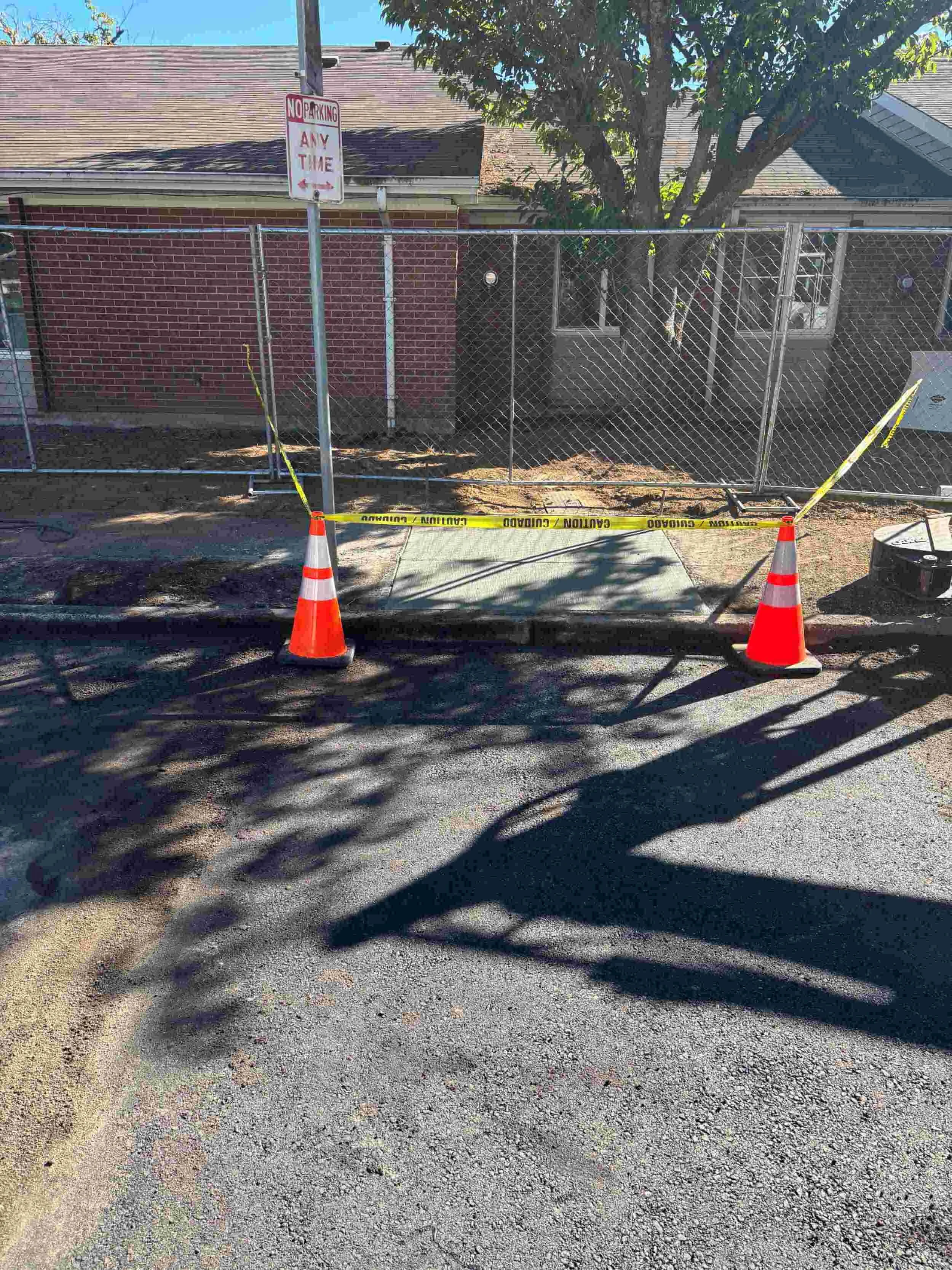A sidewalk section is cordoned off with orange traffic cones and yellow caution tape in front of a chain-link fence and a brick building with trees, under a sunny sky.