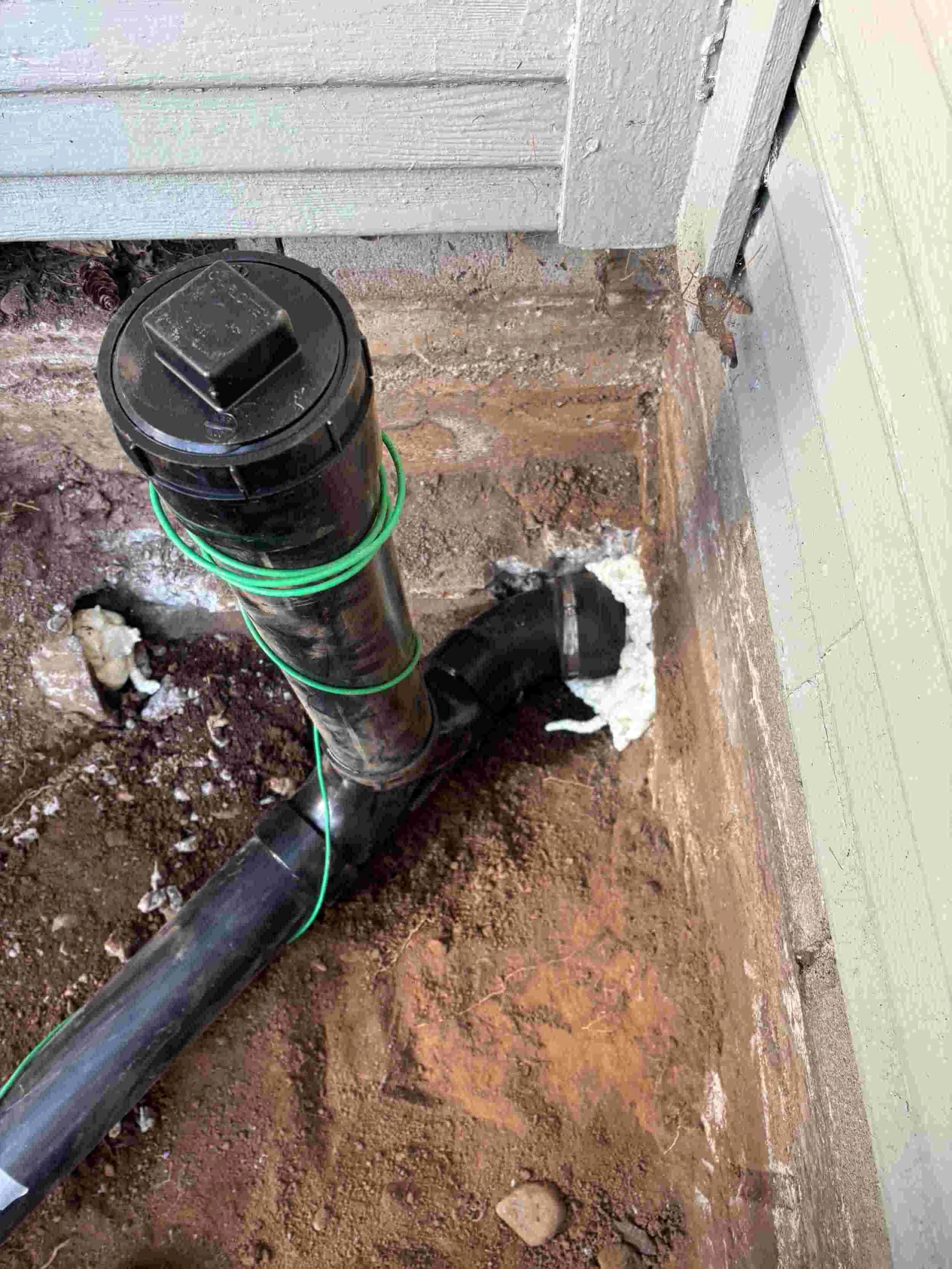 A black PVC plumbing pipe with a cap at the end, secured with green wires, runs along a dirt floor inside a building under construction or renovation, next to a wall with partially painted or unpainted surfaces.