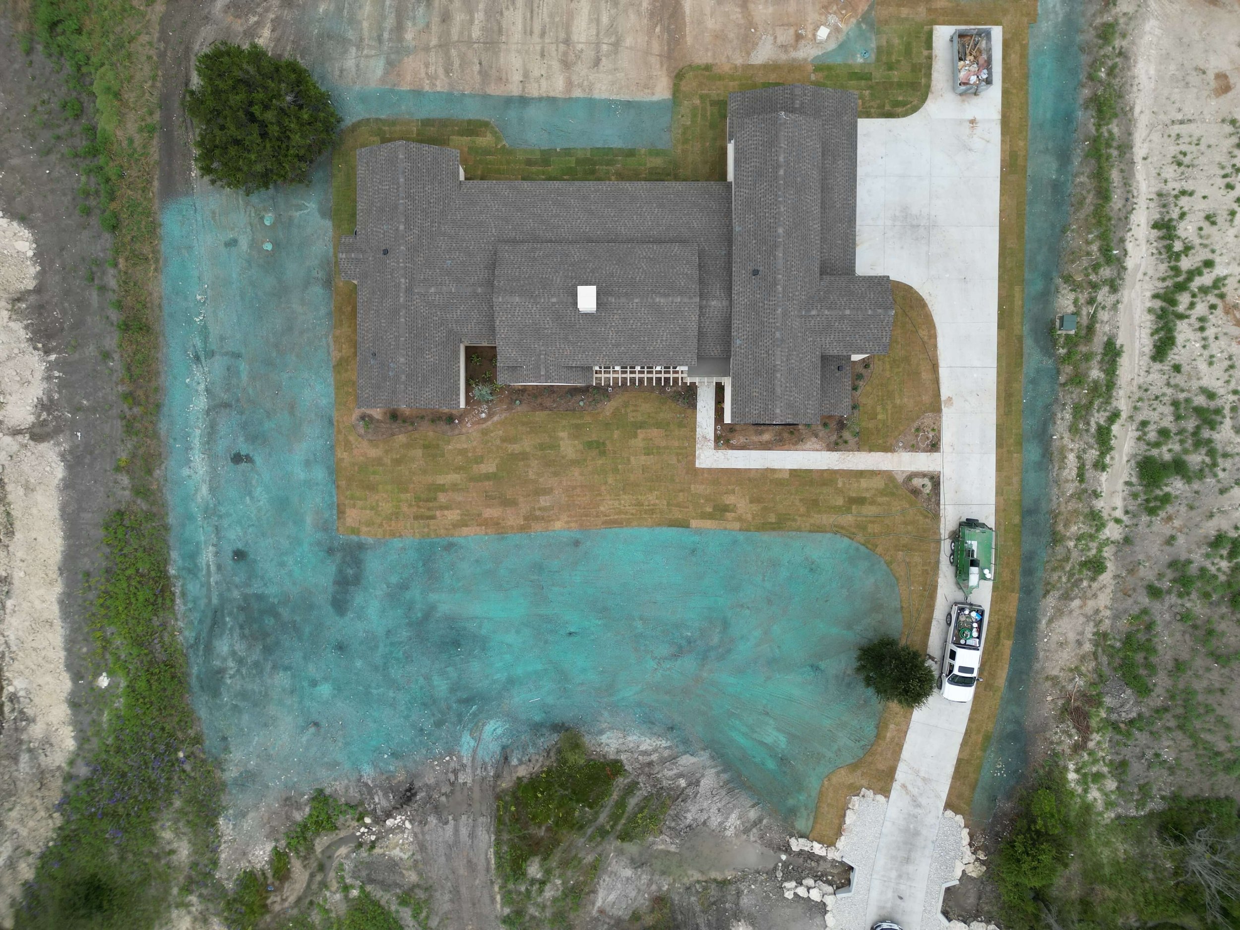 An aerial view of a house with a gray roof, surrounded by a lawn, a paved driveway with cars, and a yard with some trees and native grass or landscaping.