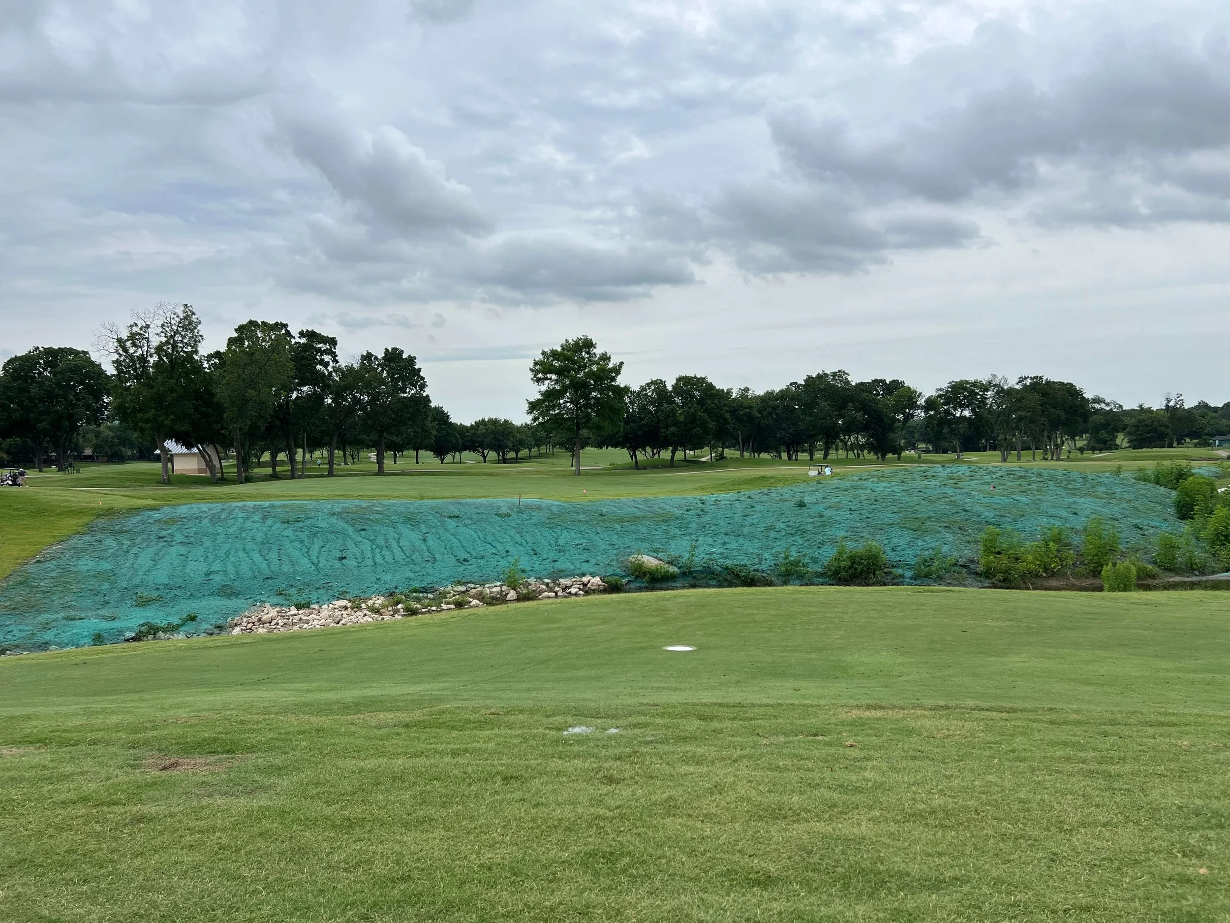 Golf course with green grass, trees in the background, a cloudy sky, and an area covered with green netting or tarpaulin, possibly for landscaping or maintenance.