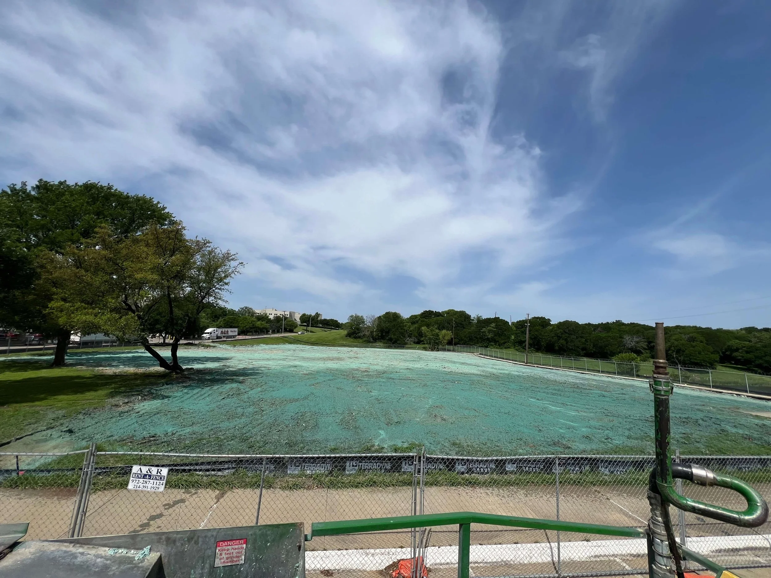 Construction site with a fenced area, green protective netting, and a water pipe in the foreground, with trees and a parking lot in the background under a partly cloudy sky.