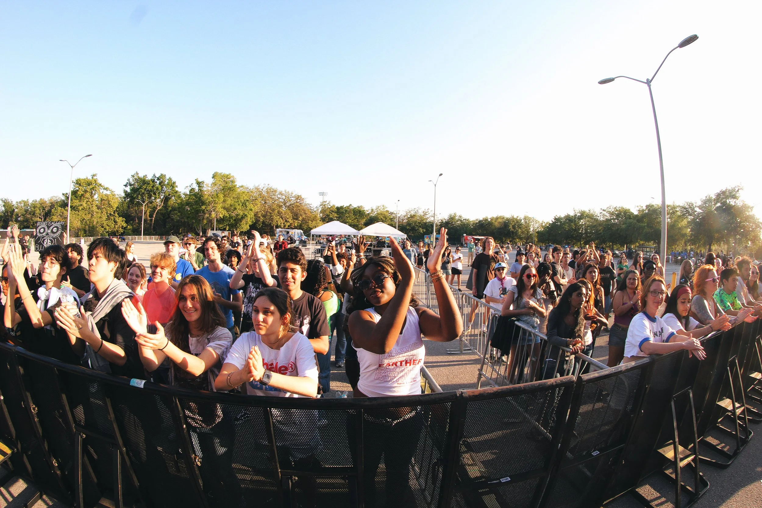 Crowd of young people at an outdoor event, standing behind barriers, some clapping and enjoying the show, with tents and trees in the background during daytime.