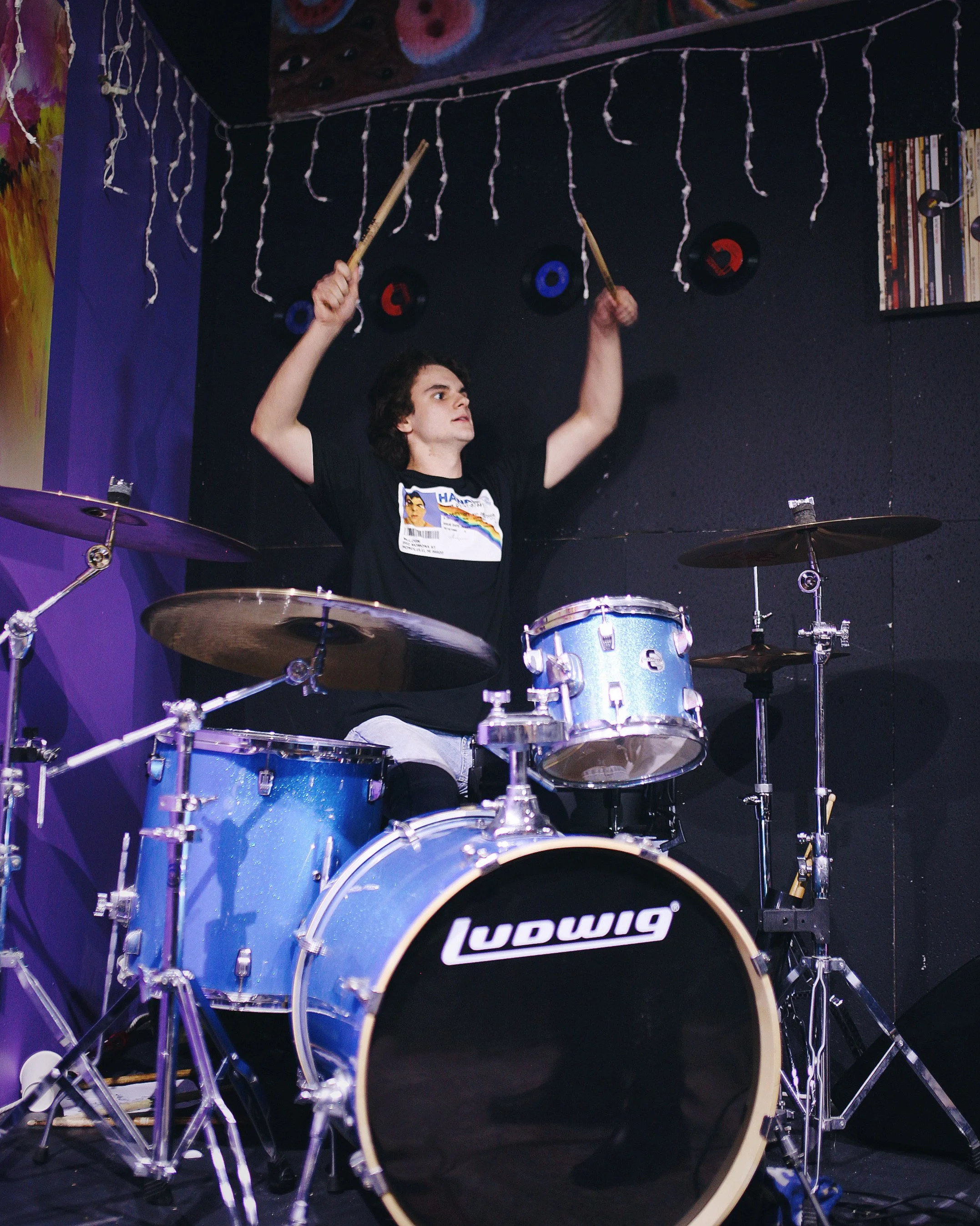 Young man playing a blue Ludwig drum set with black walls and vinyl records on the wall in the background.