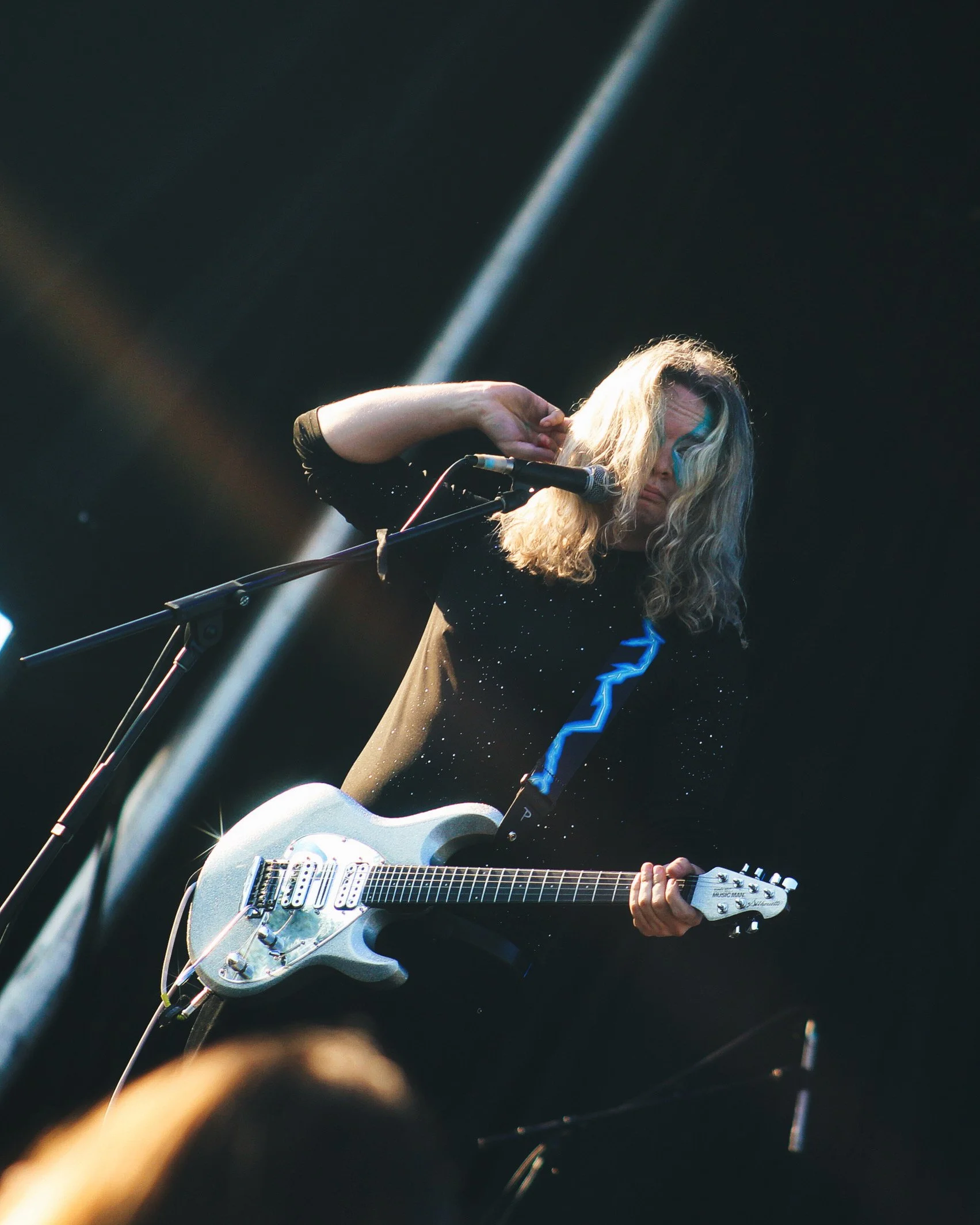 A female guitarist performing on stage, with blonde curly hair, wearing a black sparkly top, and playing an electric guitar.