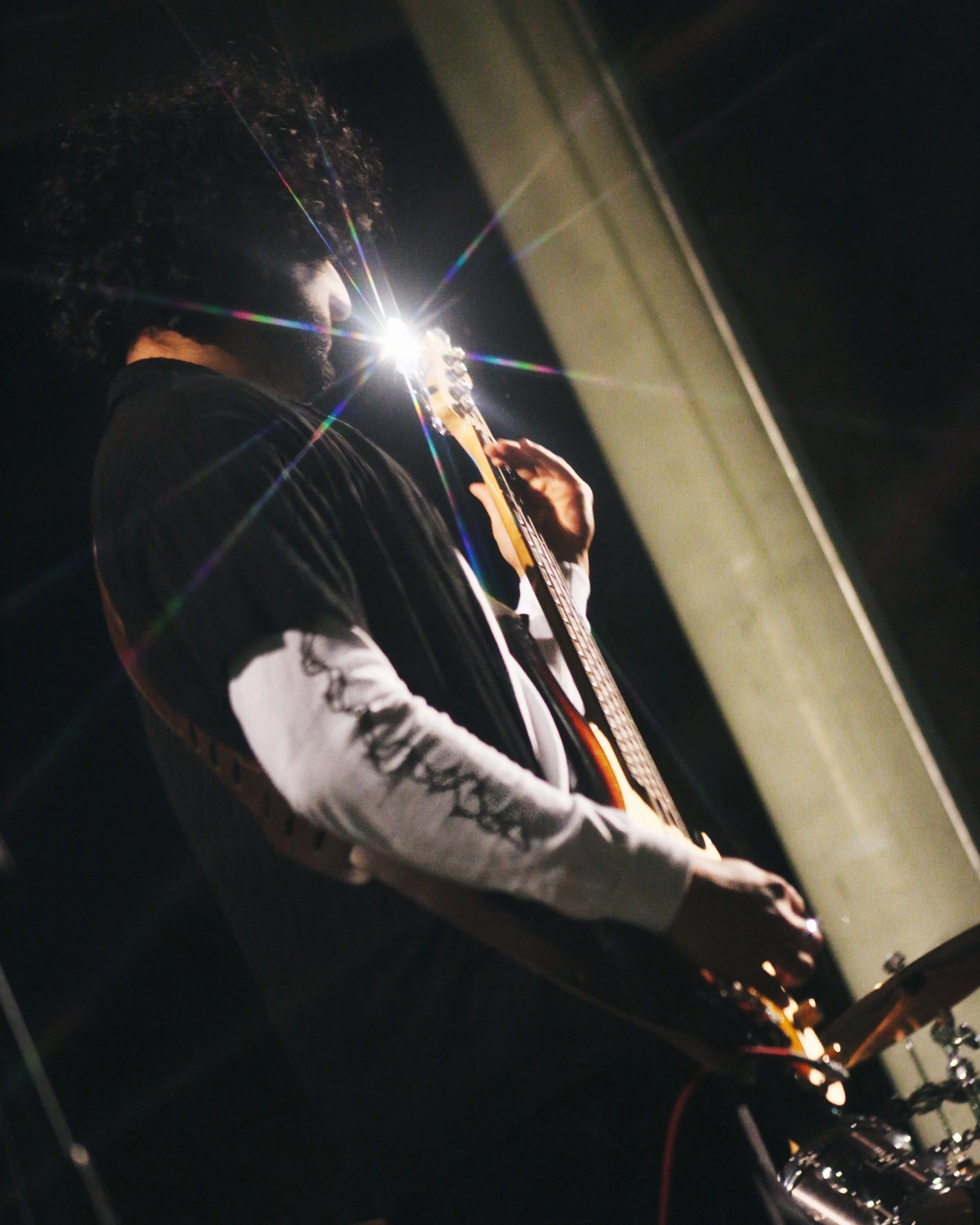 A person with curly hair playing an electric guitar in a dimly lit setting, with a bright light behind them creating a starburst effect.