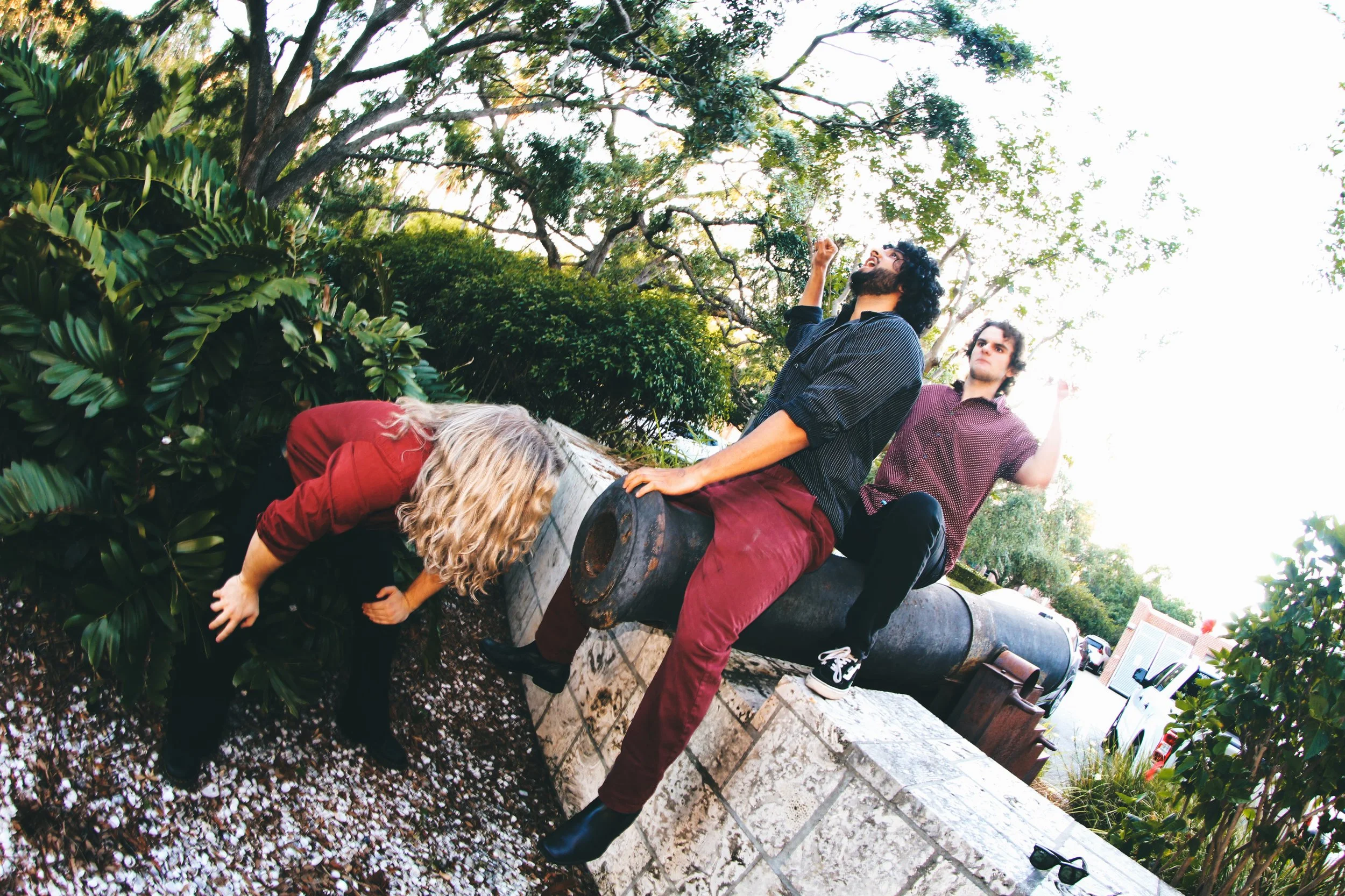 Three young people outdoors sitting and leaning on an old cannons, with trees and bushes in the background; one woman and two men, one woman with blonde hair measuring water on her hand, the man with black curly hair and beard with one hand up, and a