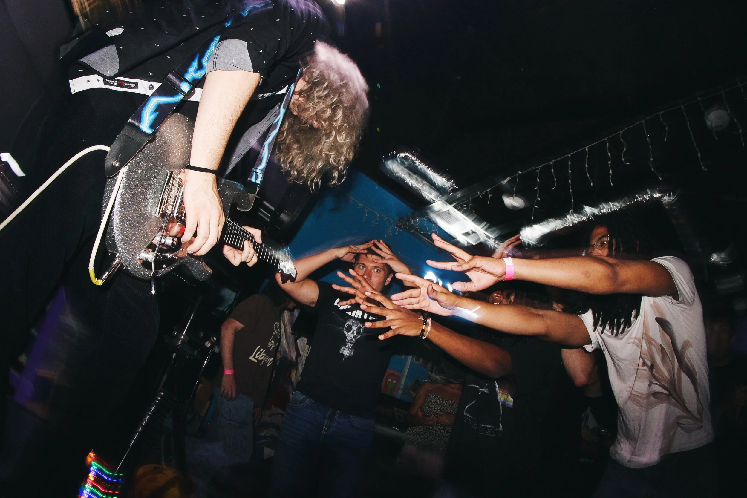 A musician with curly hair playing an electric guitar on stage with an audience reaching out to him.