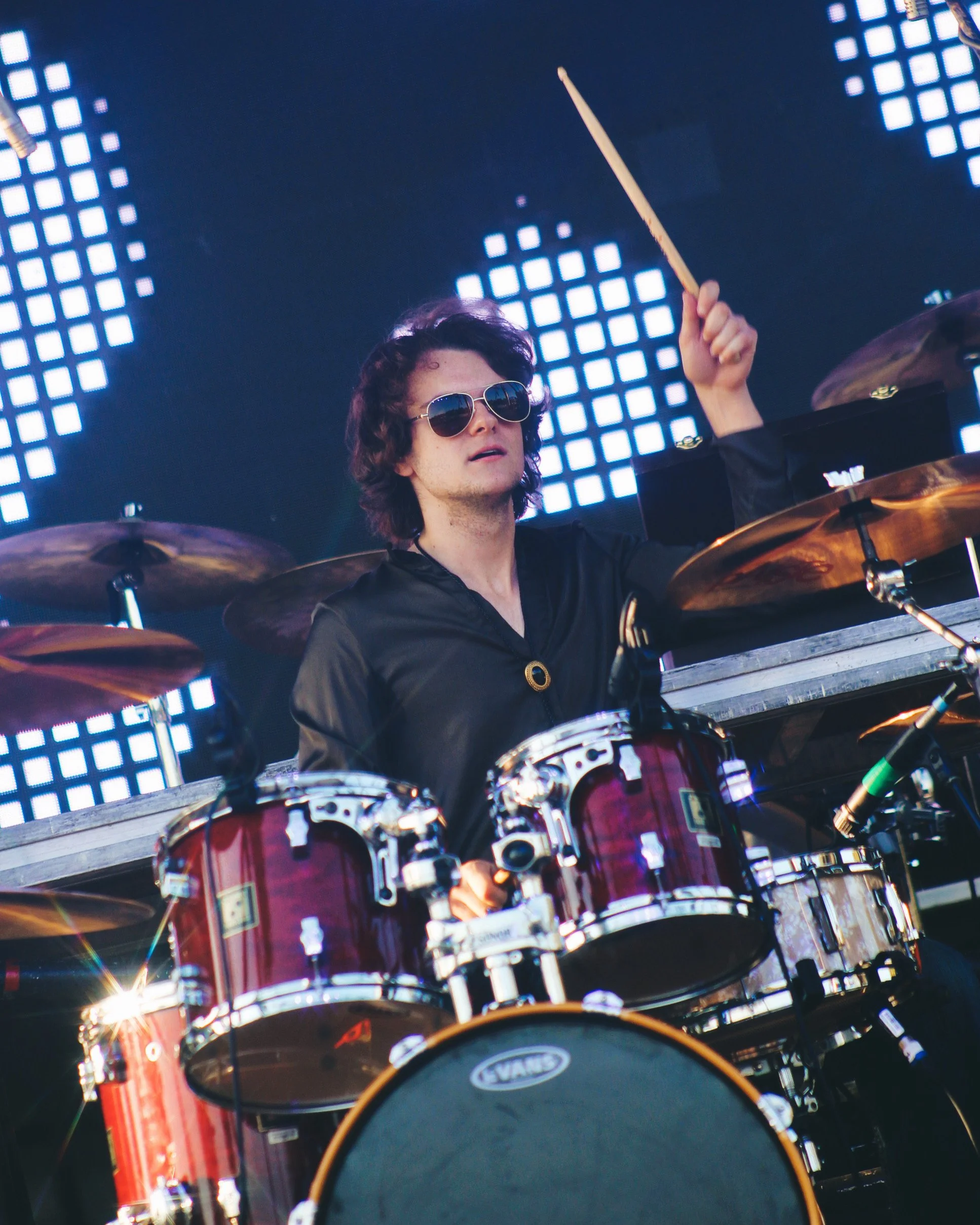 A male drummer wearing sunglasses playing a drum set during a concert, with a digital LED background display.
