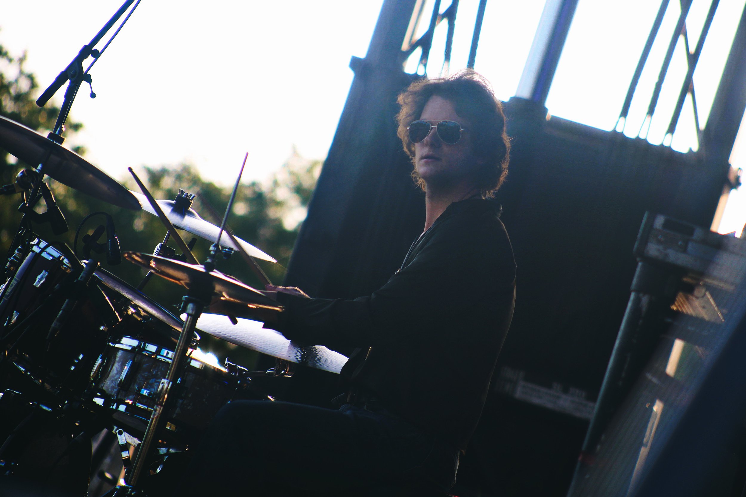 A man with sunglasses sitting at a drum set outdoors during sunset