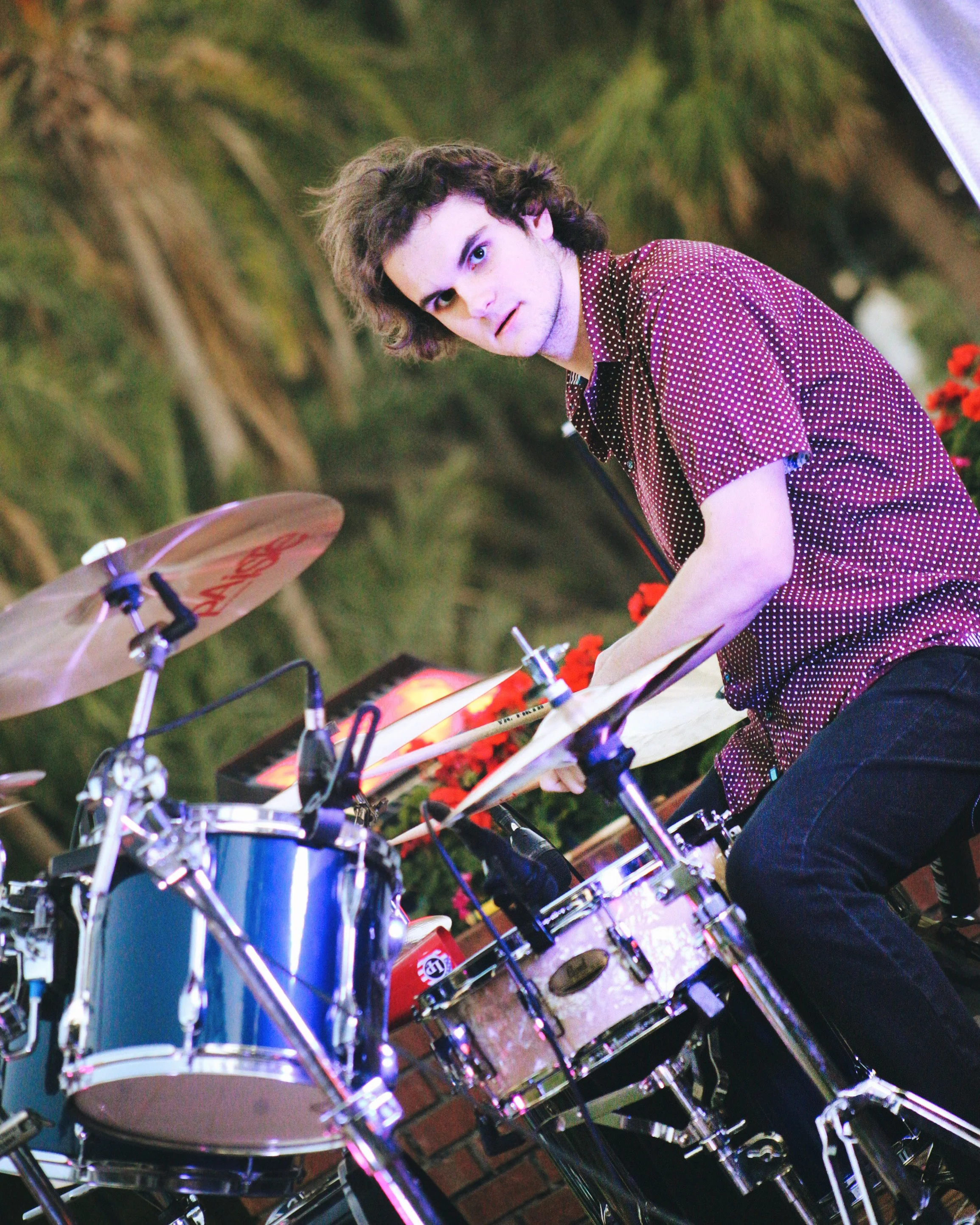 A young man with curly hair playing a drum set outdoors at night, with green foliage in the background and red flowers nearby.