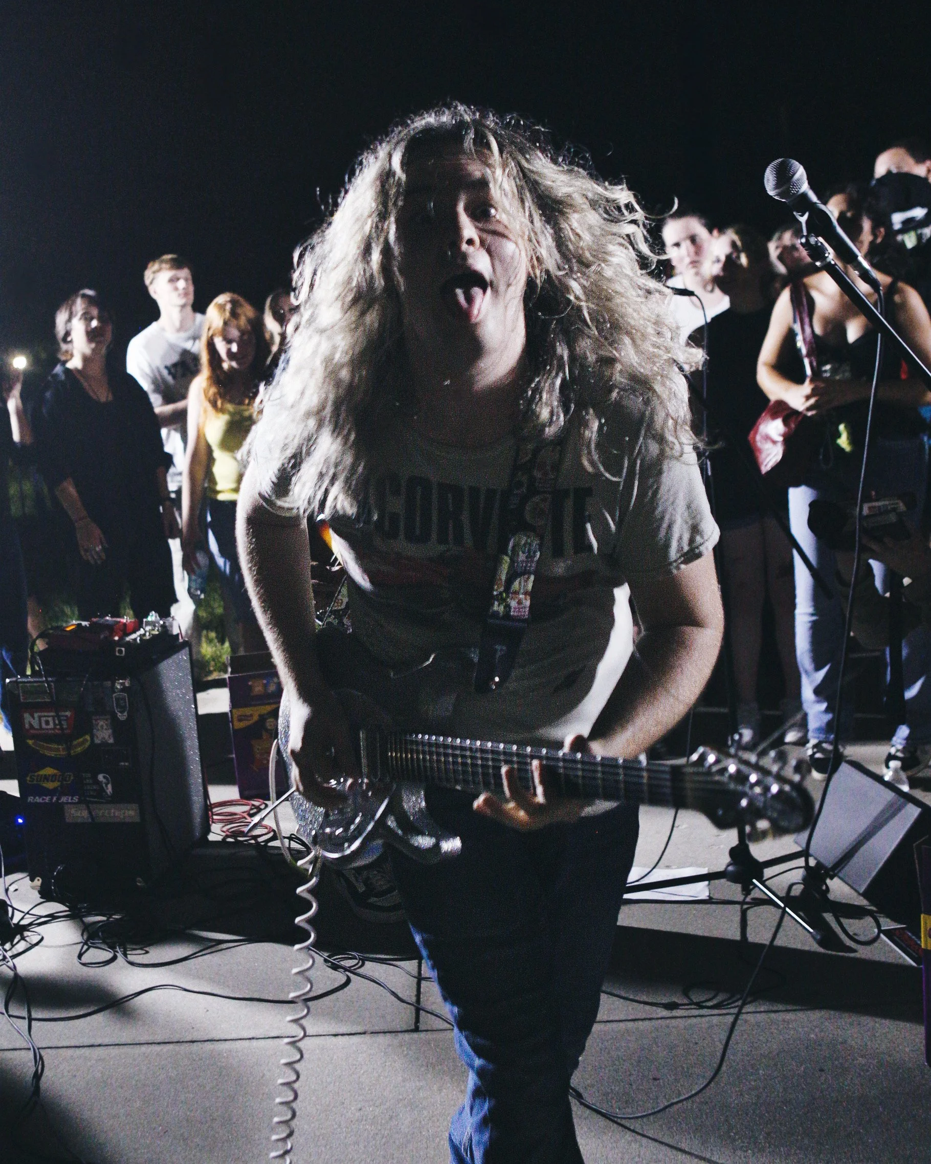 A woman with long, curly blonde hair playing an electric guitar during a nighttime outdoor concert, with a crowd of people standing behind her.
