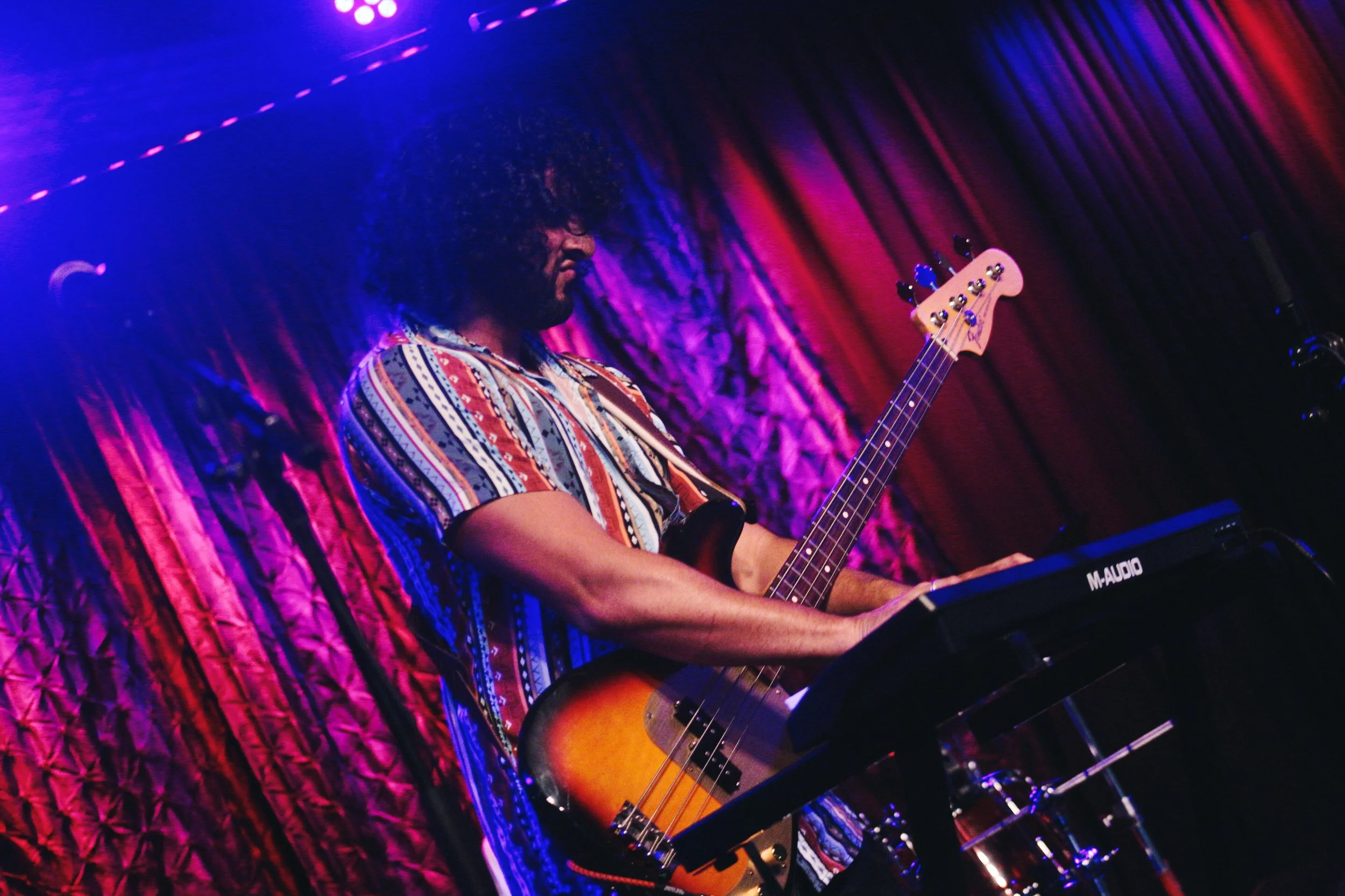 A musician playing a bass guitar on stage under colorful purple and red lights, with a textured purple curtain in the background.