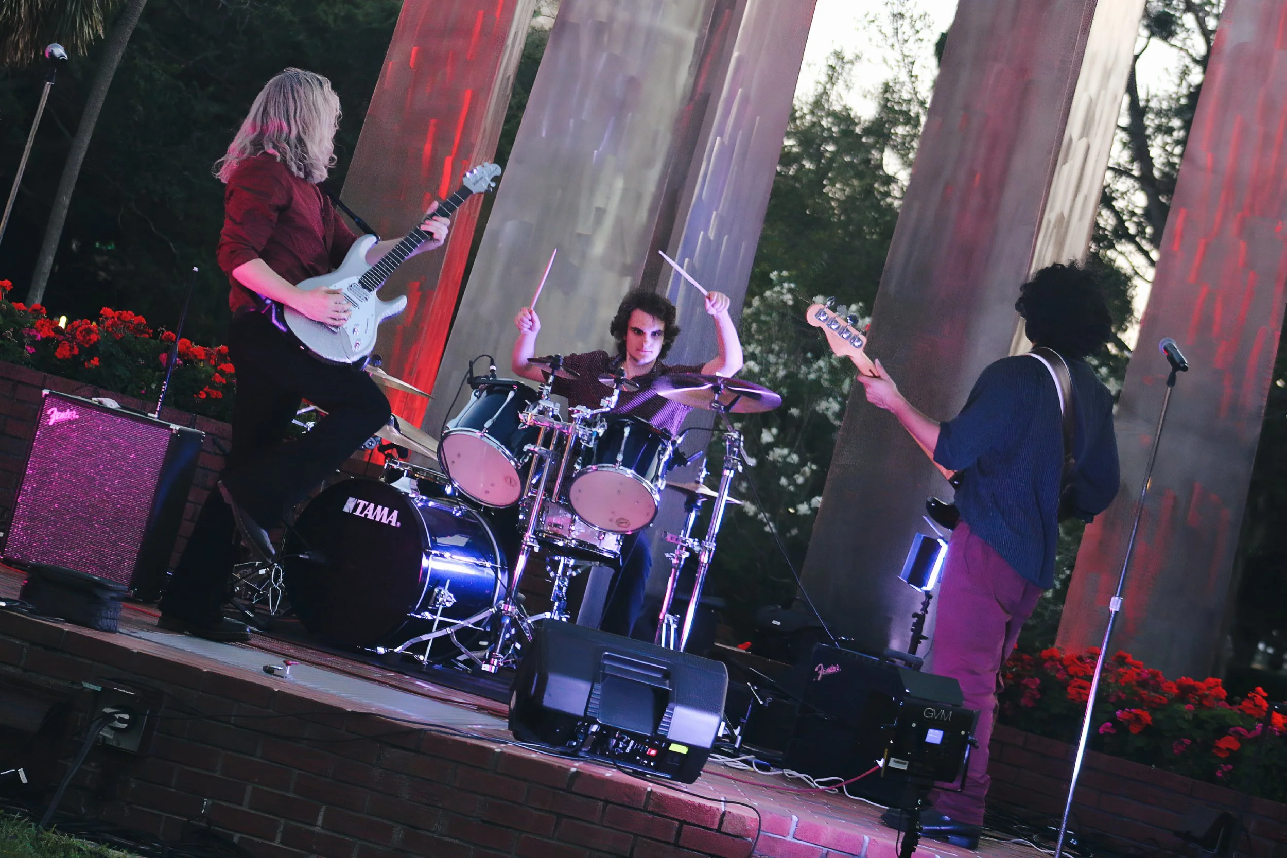 A band performing live outdoor concert, with a guitarist, a drummer, and a bassist on stage with amplifiers and microphones, surrounded by trees and decorative flowers.