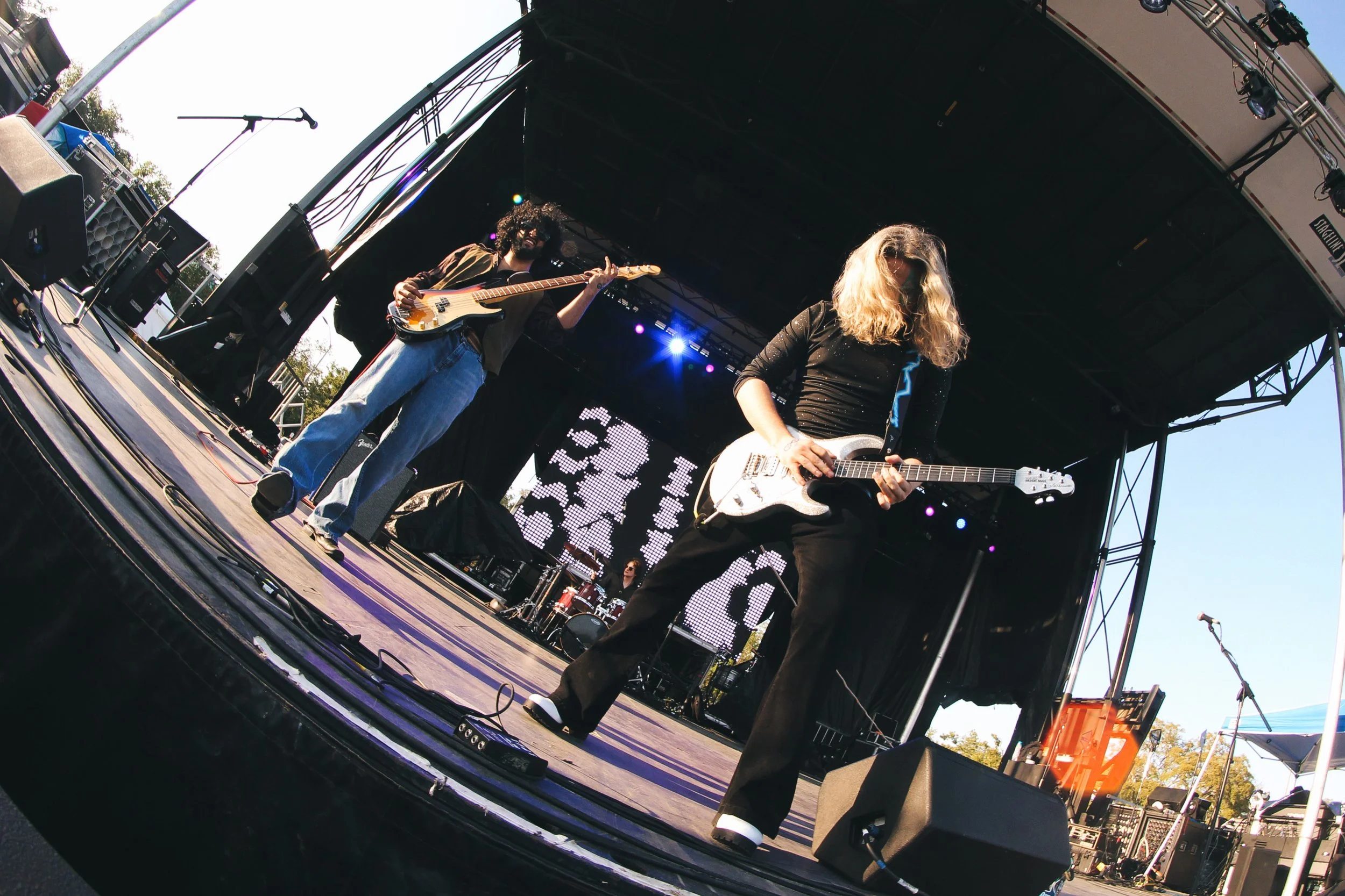Two female musicians performing on an outdoor stage with guitars, one playing bass and the other playing an electric guitar, with a drummer visible in the background and a large screen behind them.