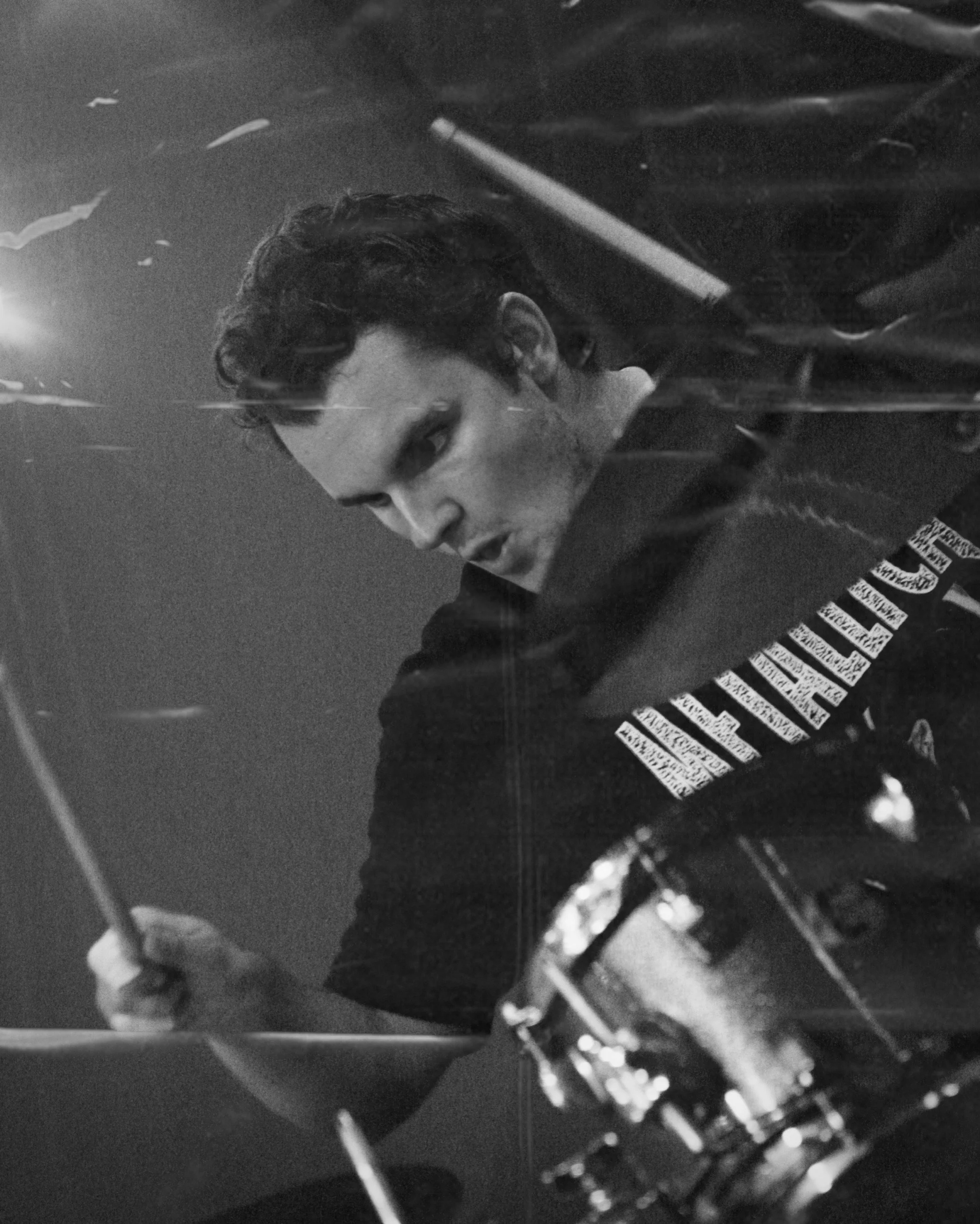 A black and white photo of a man playing piano with a drum set nearby, wearing a T-shirt with a message, captured from an angled perspective.
