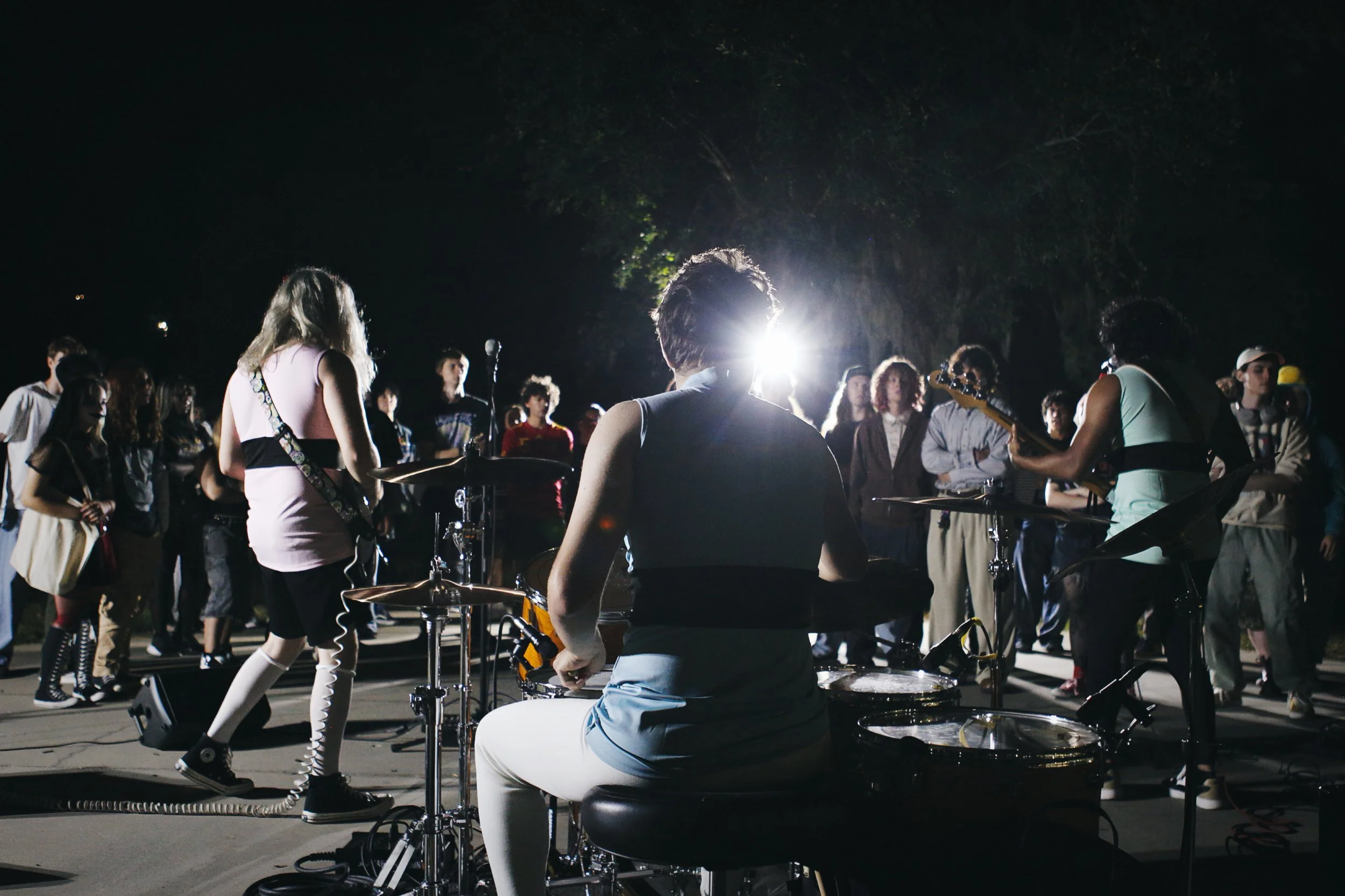 A live outdoor music performance at night, with a band playing and an audience gathered around.