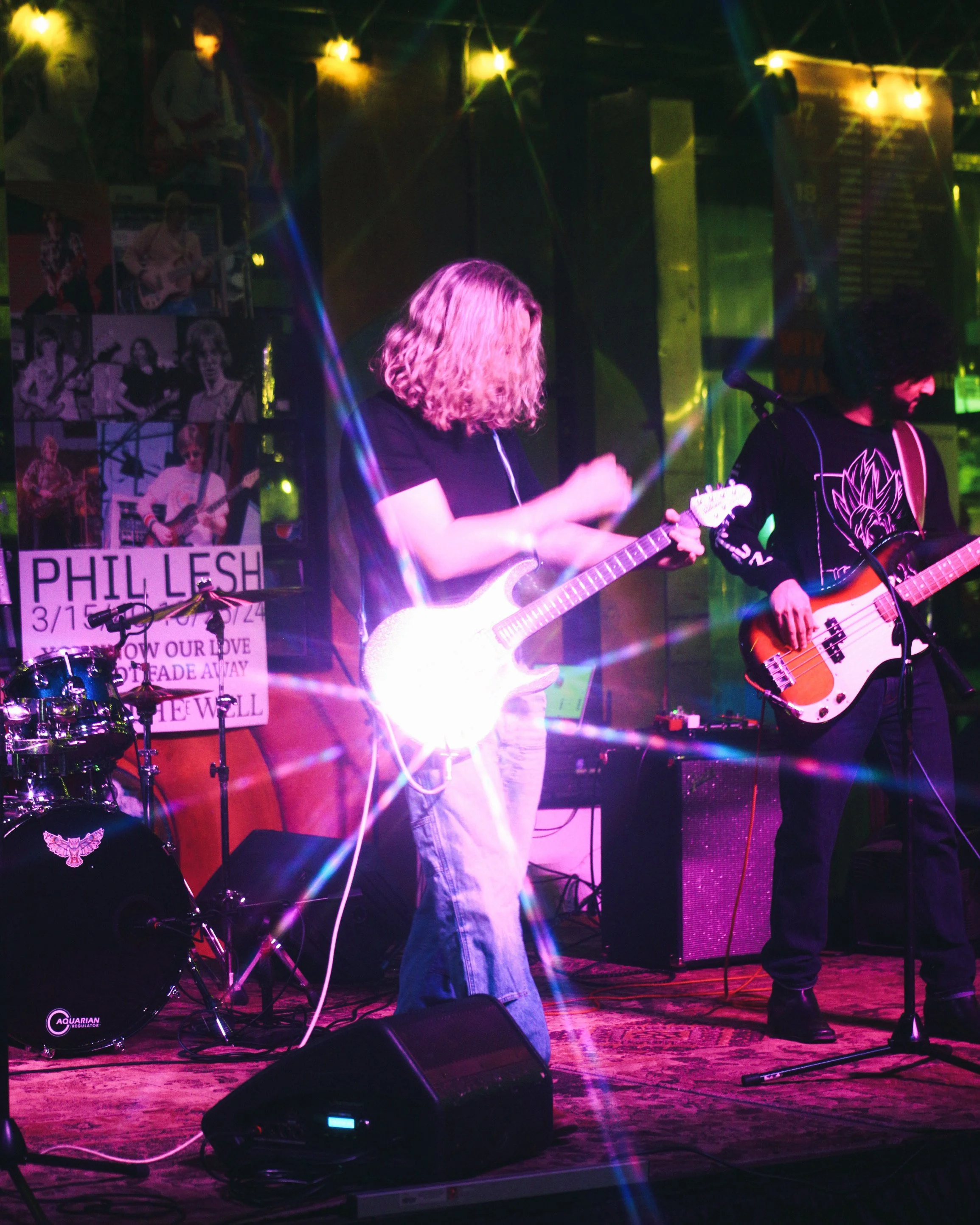 Musicians performing on stage with guitars and drums, colorful stage lighting, posters on the wall, and a banner for Phil Lesh.