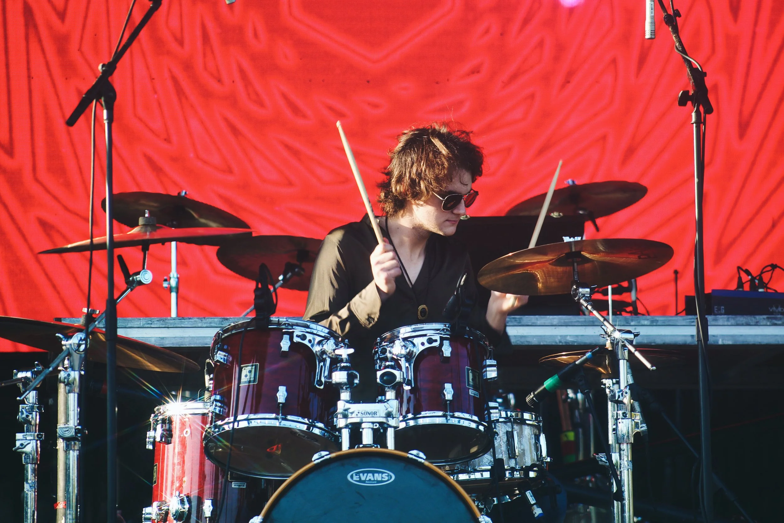 A young man wearing sunglasses, a black shirt, and a tan jacket playing a red drum set on stage with a bright red textured backdrop.