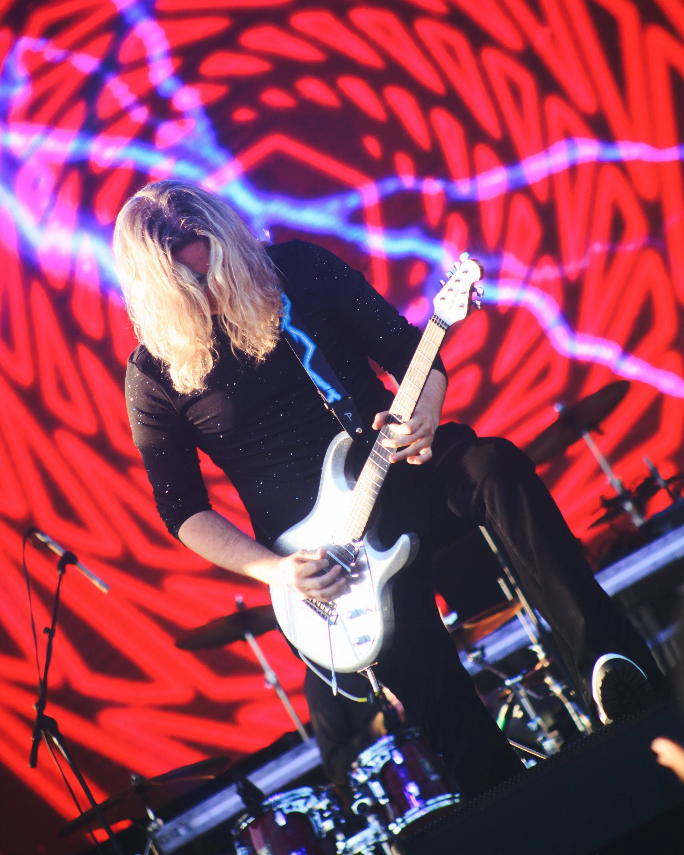 A musician with long blonde hair plays an electric guitar on stage in front of a red and black swirling background.
