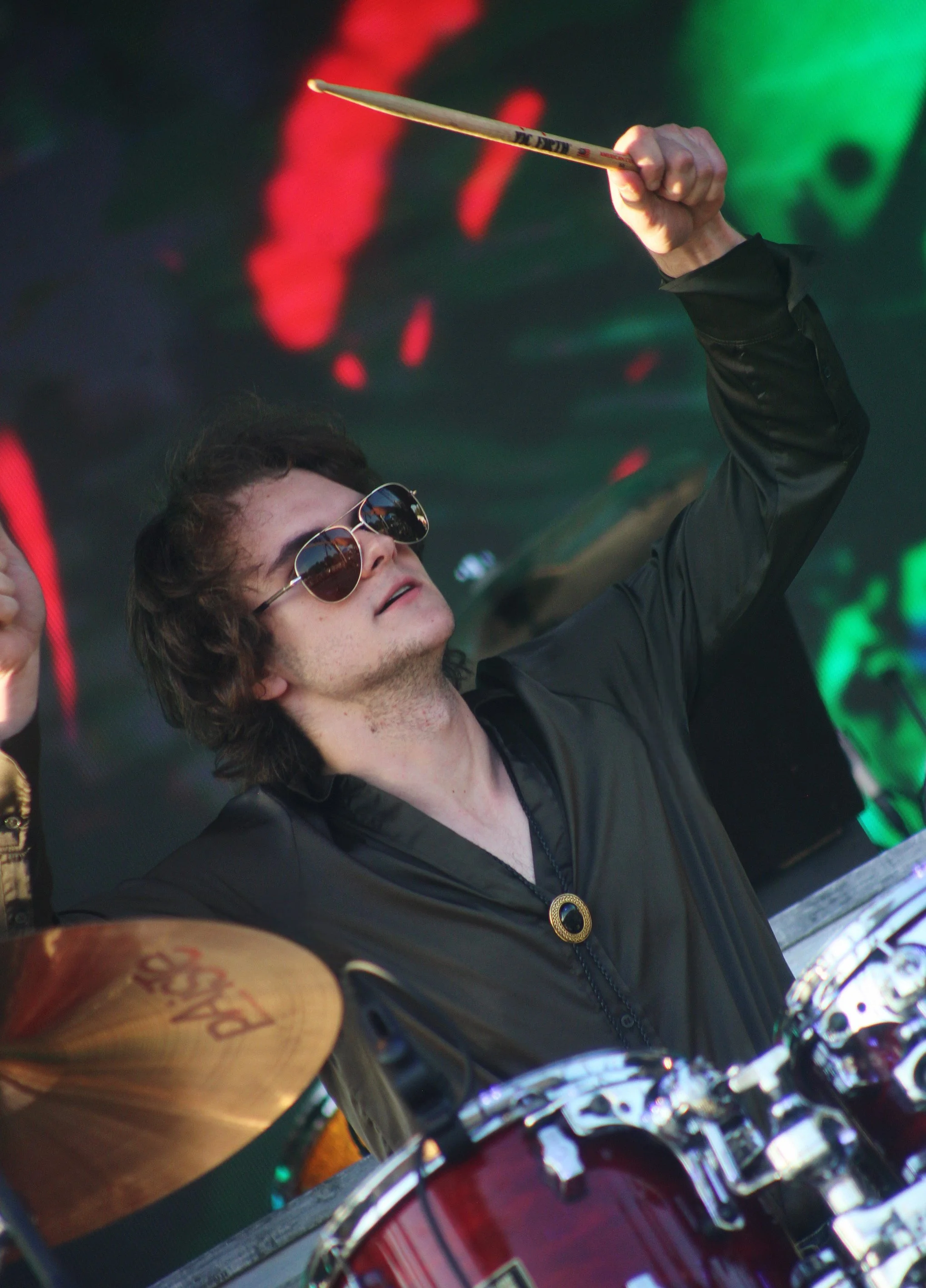 A young male musician with long, curly hair, sunglasses, and a black jacket playing drums on stage, holding a drumstick in the air.