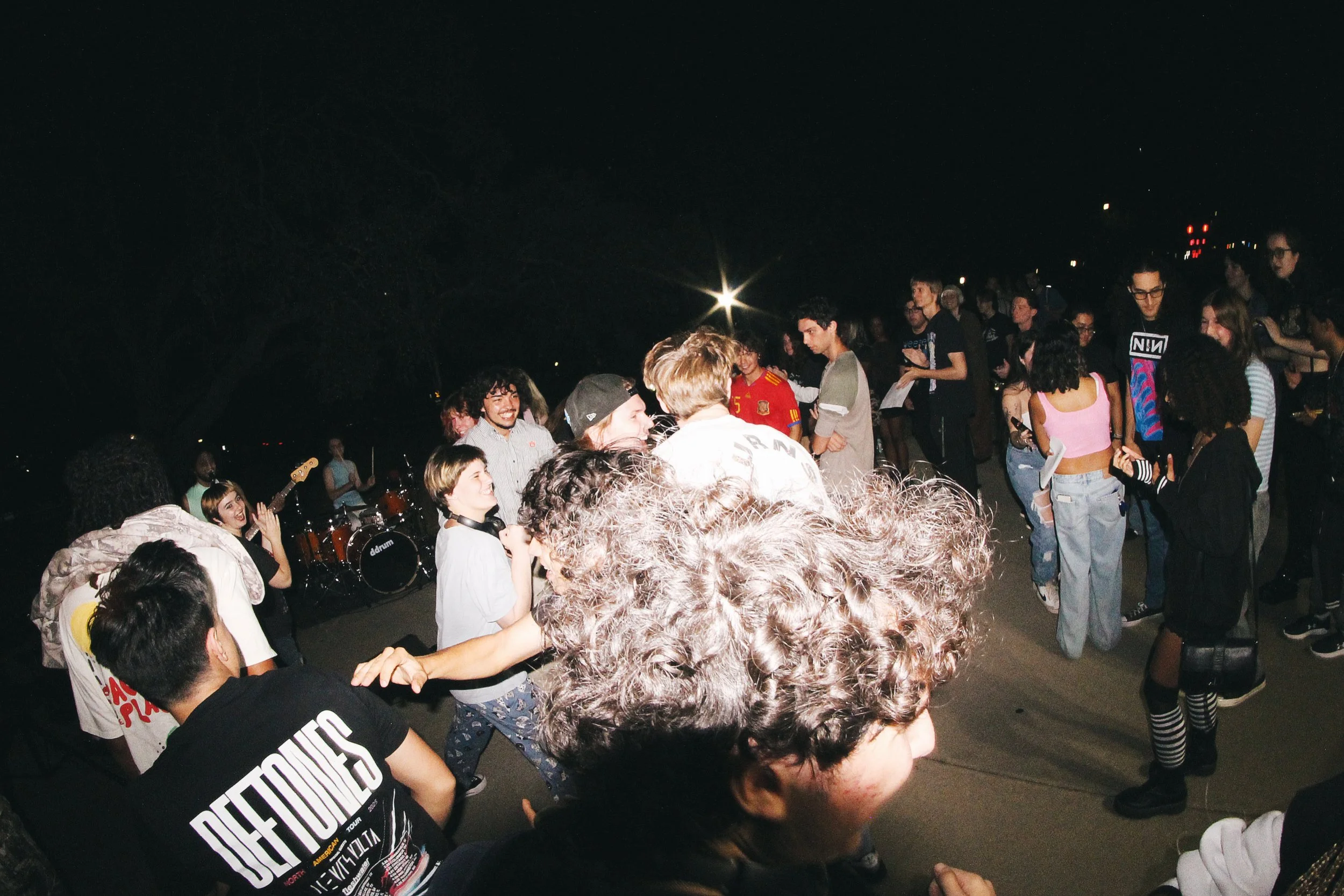Group of young people dancing and socializing outdoors at night, with a live band playing in the background.