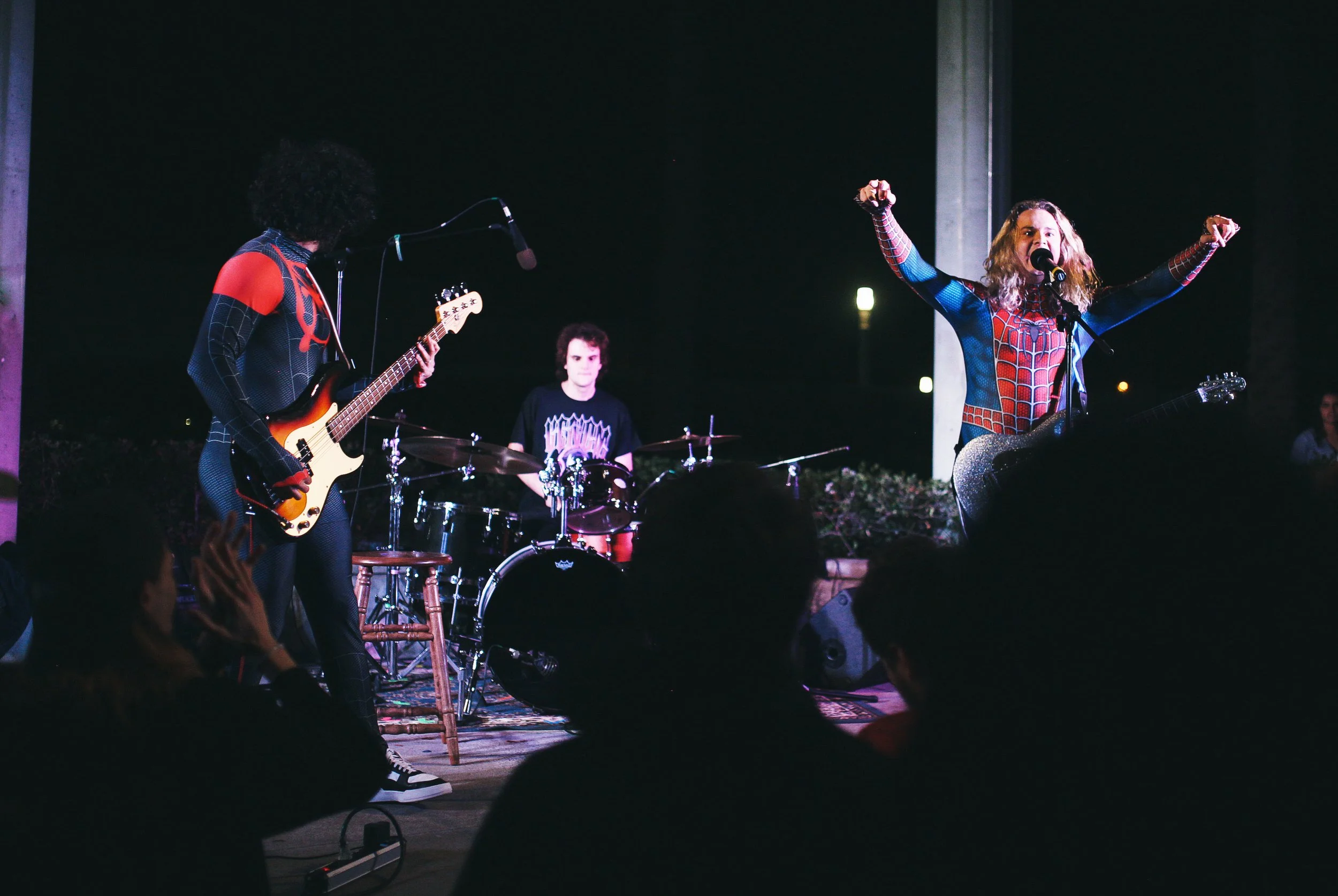 A band performing on stage with a guitarist in a Spider-Man costume, a drummer, and a bassist, at a concert with audience silhouettes in the foreground.