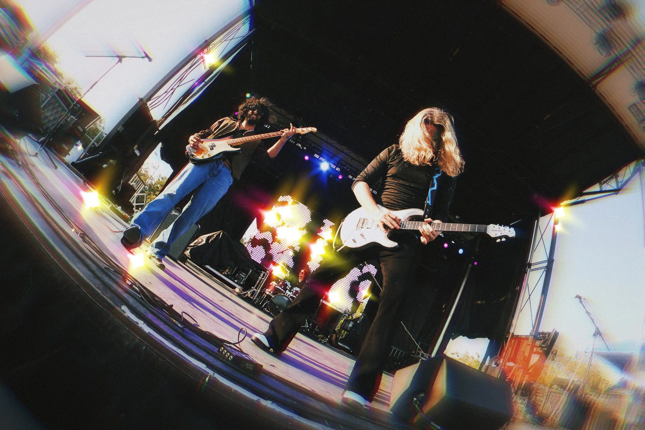 Two female musicians playing electric guitars on an outdoor stage with colorful stage lights and a large digital display in the background.