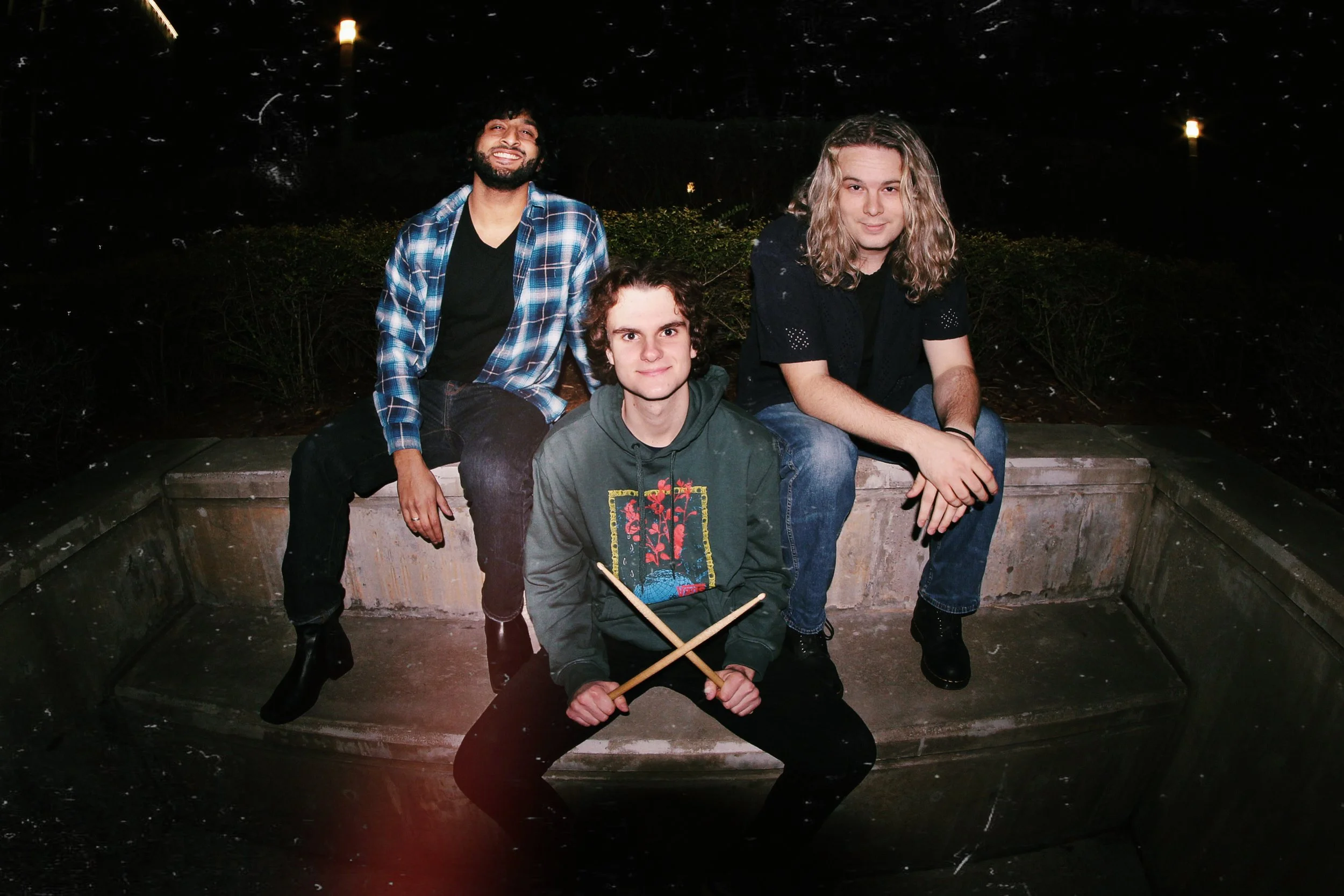 Four young men sitting on a concrete bench outdoors at night, with three of them smiling and one holding drumsticks, during a nighttime gathering.