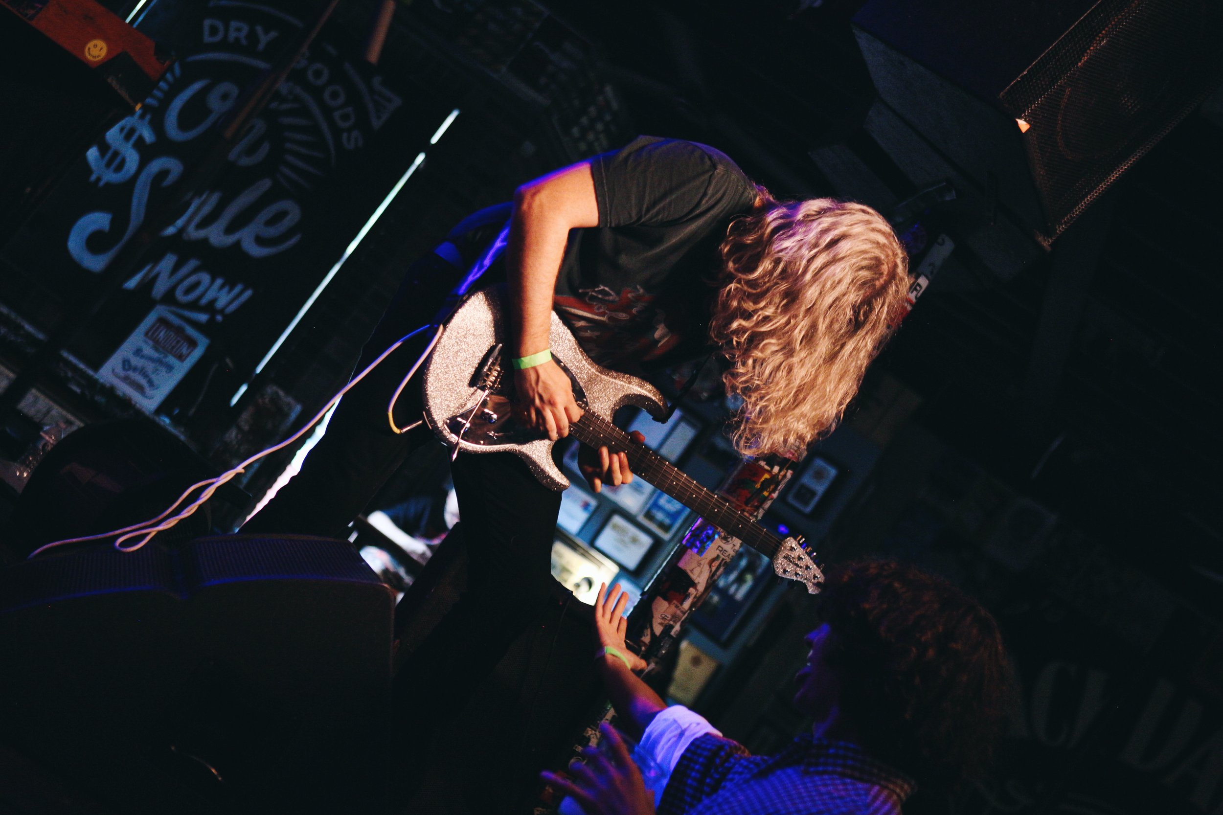 A musician with curly blonde hair playing an electric guitar on stage, with another person sitting in front of the stage reaching out.