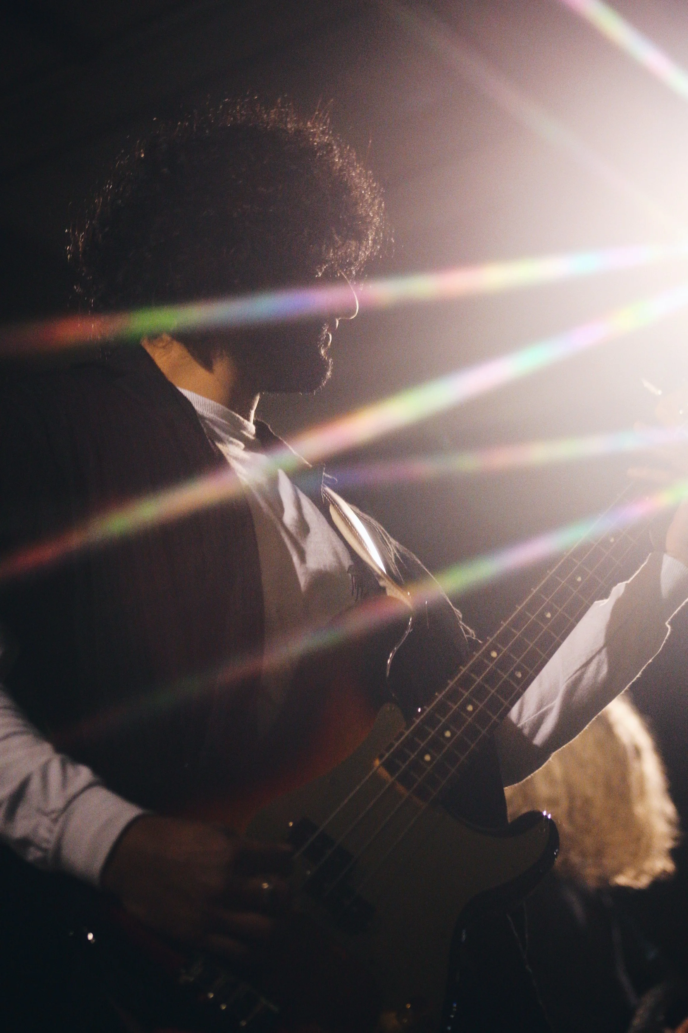 A man with curly hair and a beard playing an electric bass guitar, illuminated by dramatic stage lighting with lens flare effects.