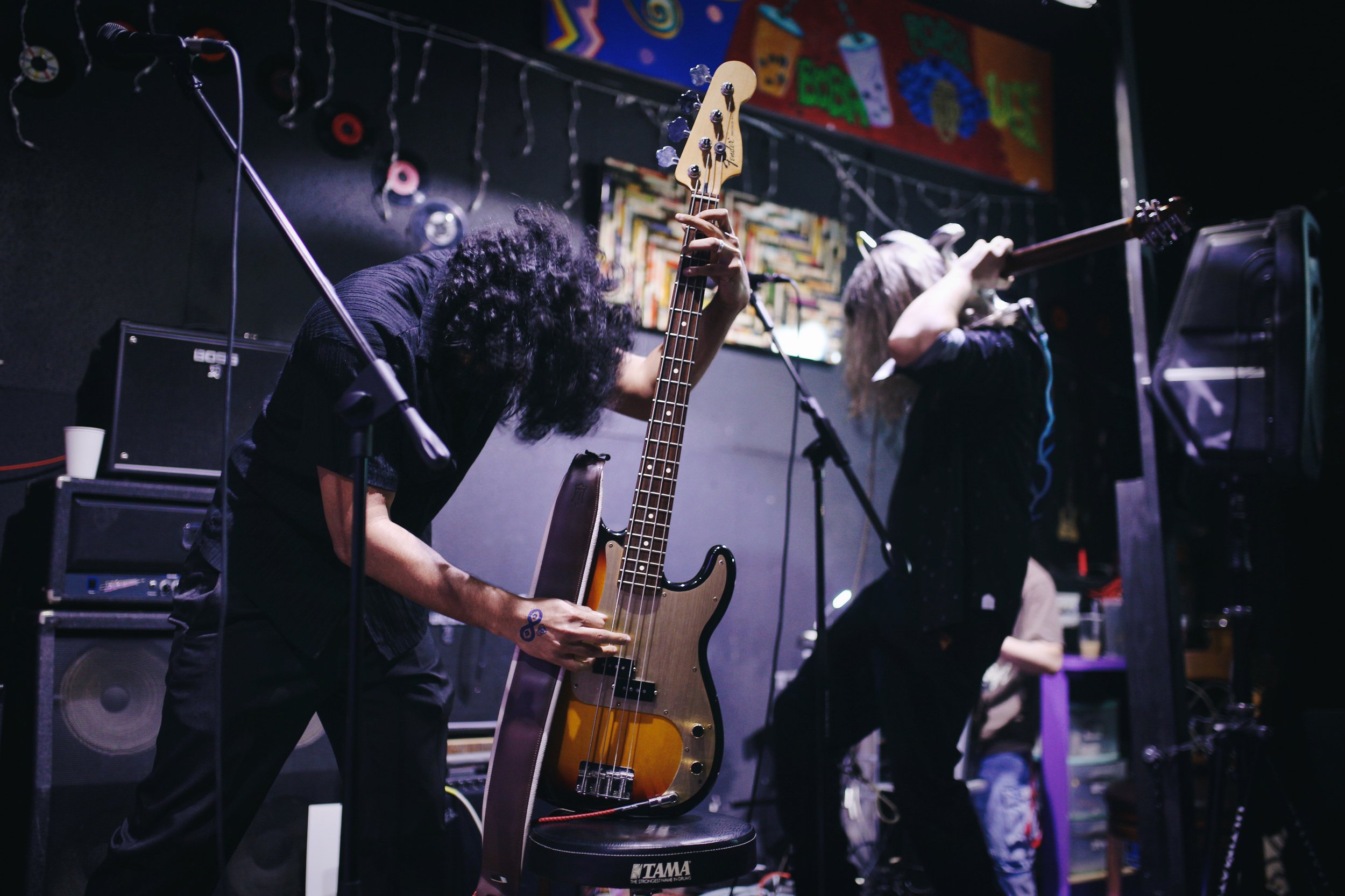 Two musicians performing with guitars on a stage with colorful artwork and string lights in the background.