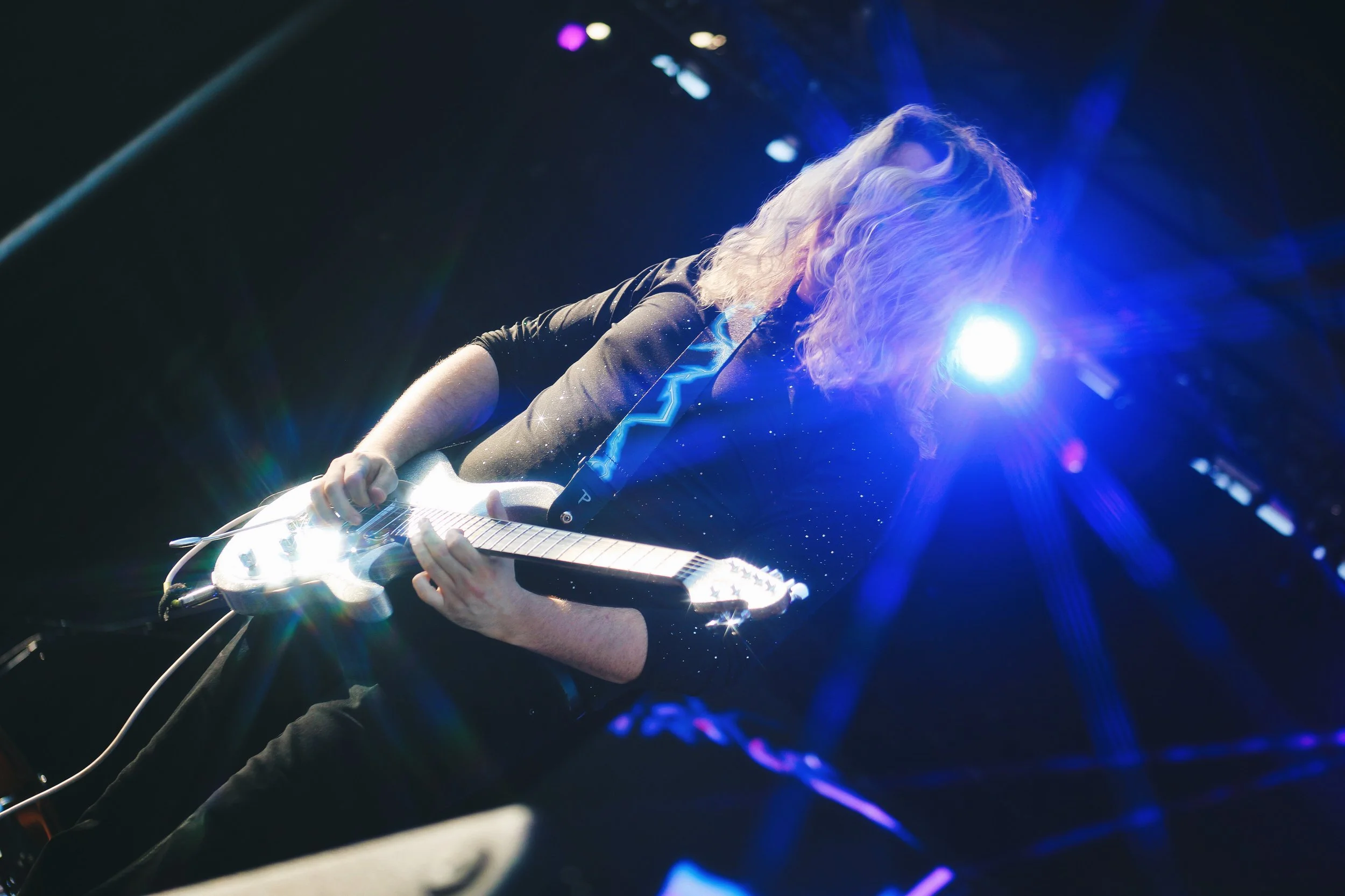 A woman with curly blonde hair playing an electric guitar on stage, illuminated by bright blue stage lighting.