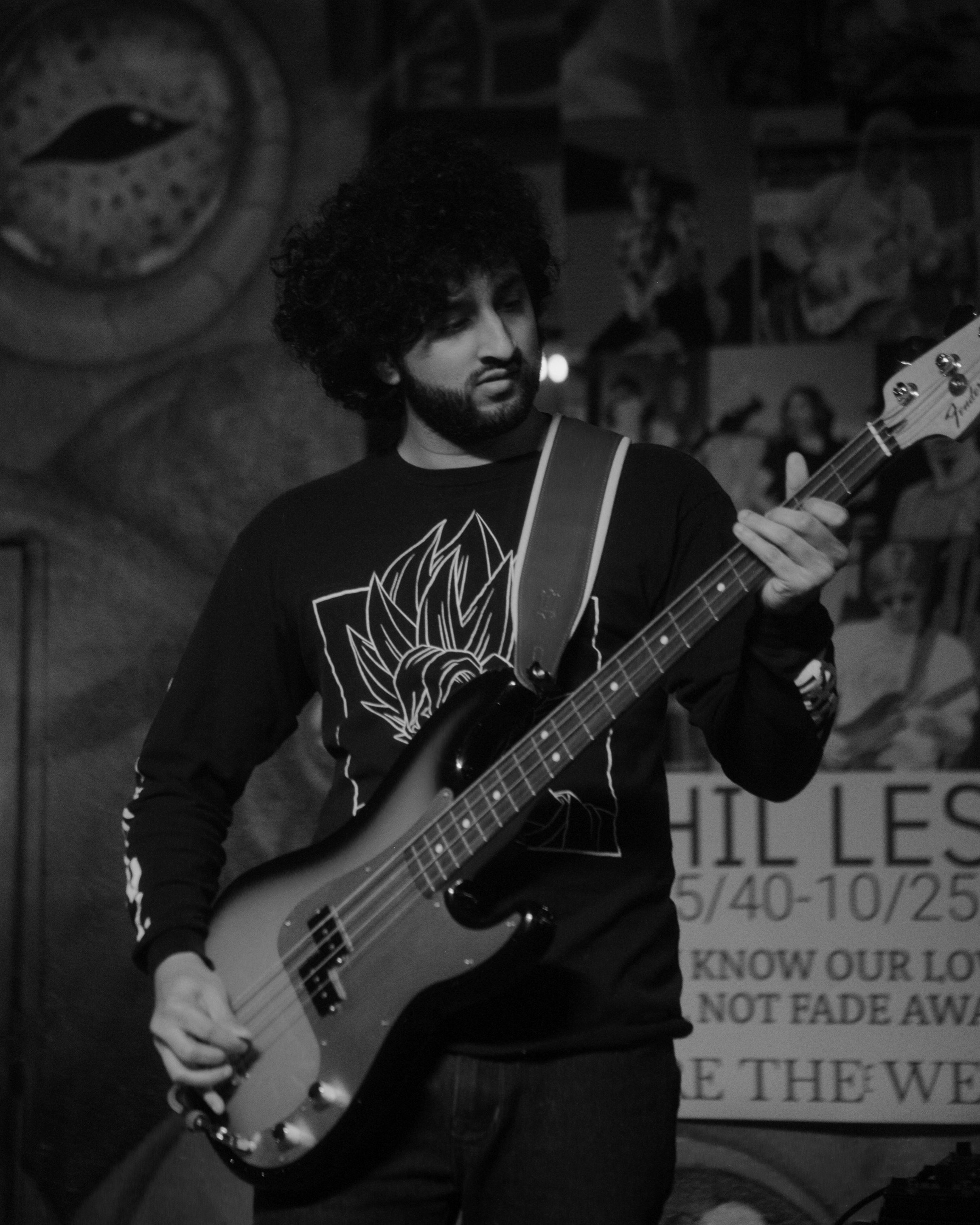 A young man with curly hair and a beard playing an electric bass guitar in a dimly lit venue, with posters on the wall behind him.