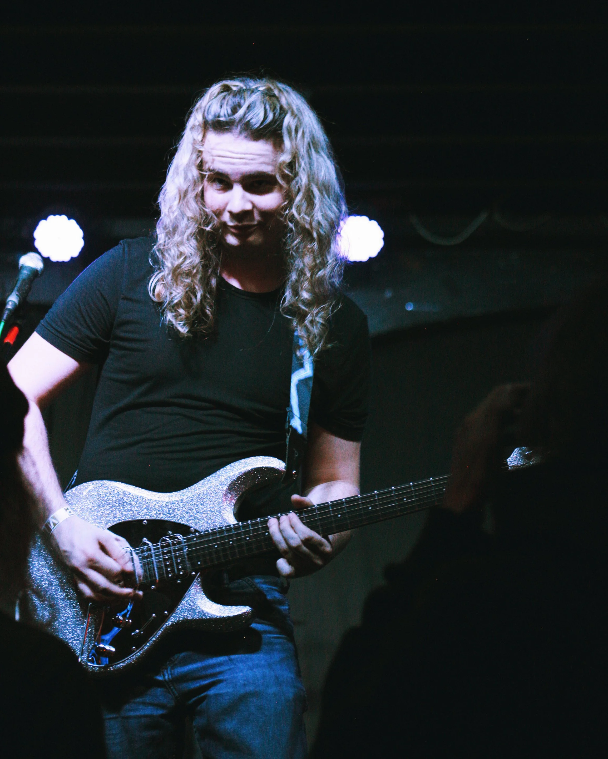 A guitarist with long, curly blonde hair playing an electric guitar on stage under spotlights.