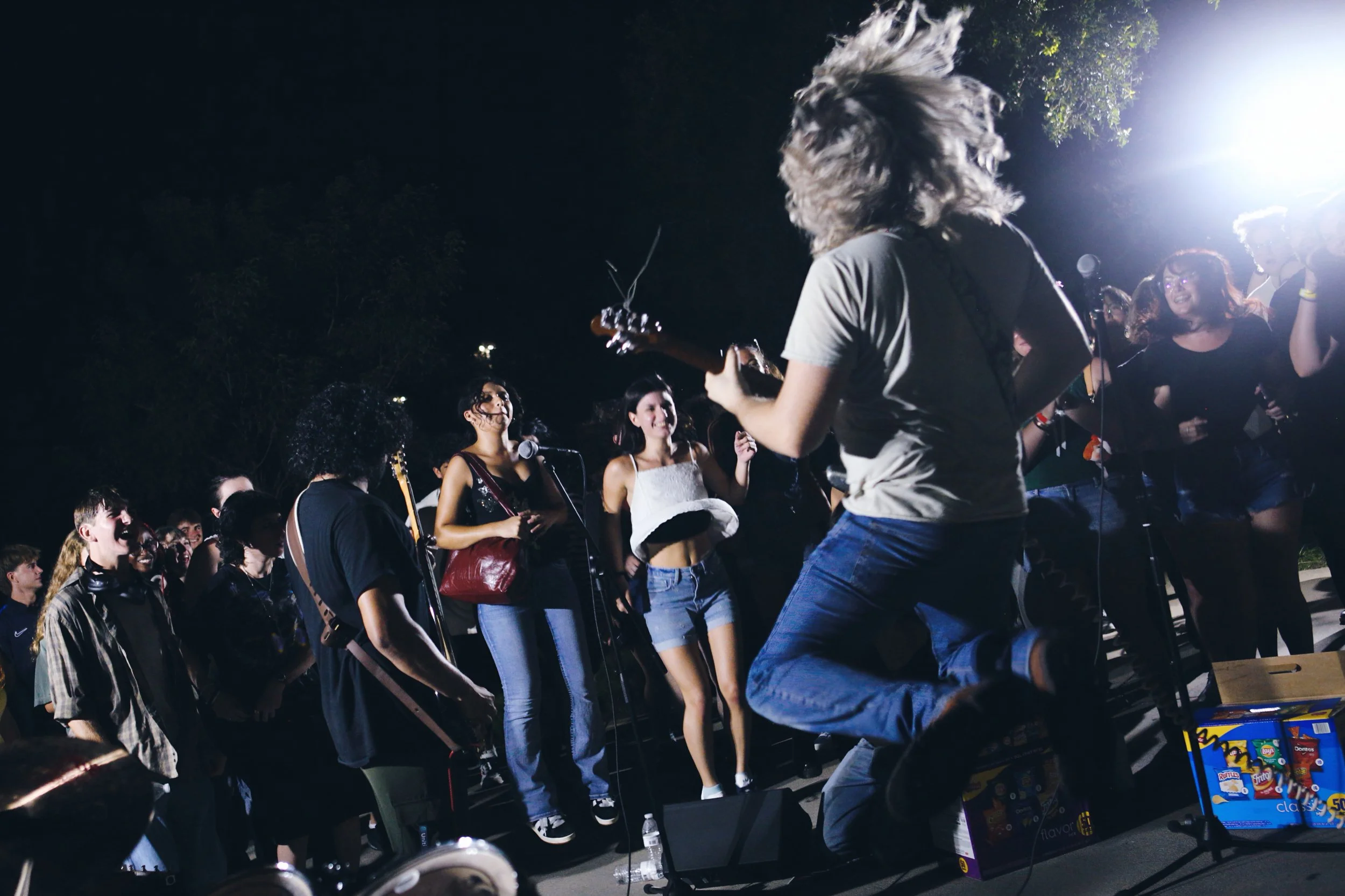 A musician with long hair playing an electric guitar at an outdoor concert at night, surrounded by a smiling audience enjoying the performance.