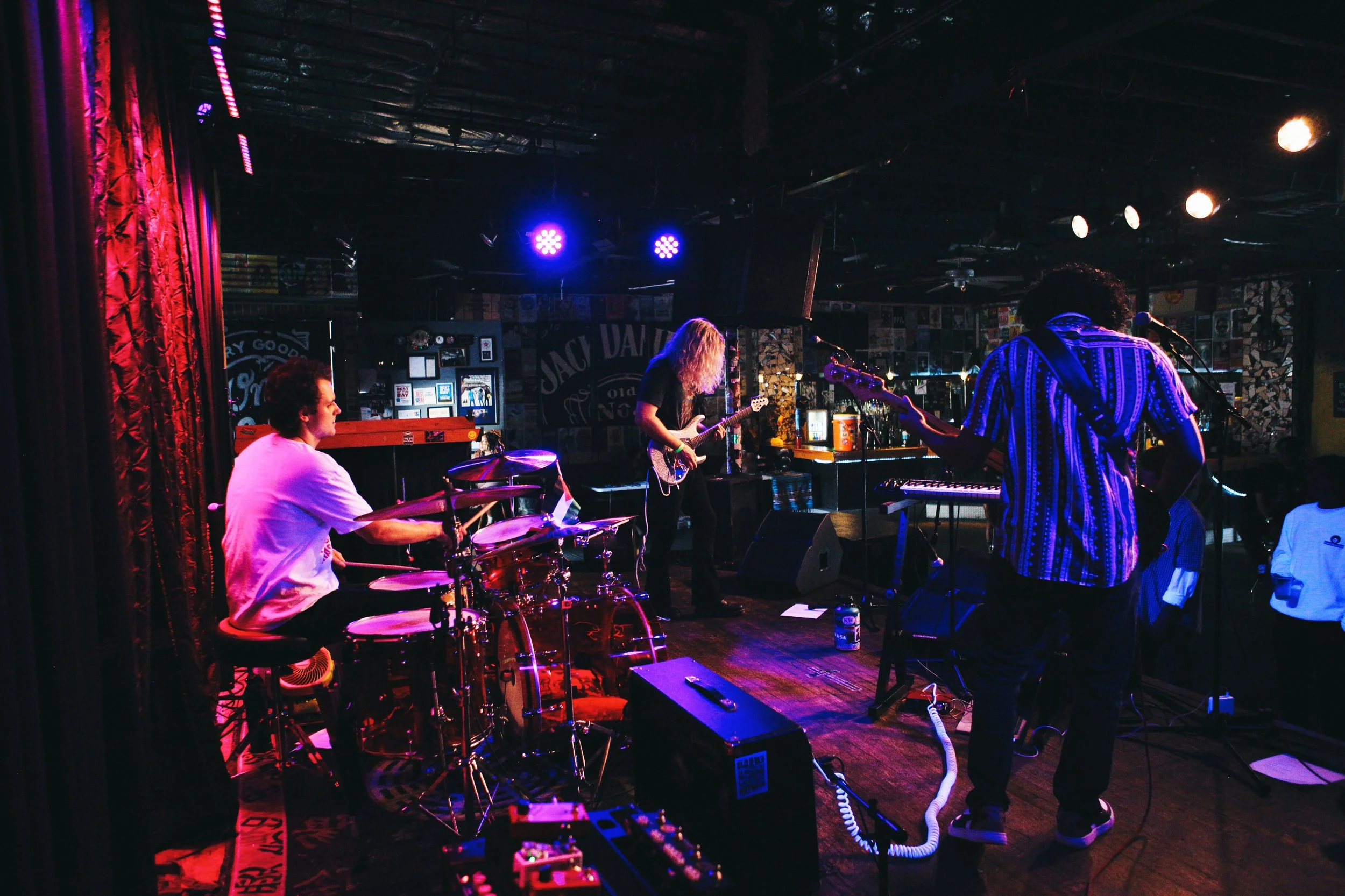 A band performing live on stage at a bar with purple and orange lighting; a drummer on the left, a guitarist beside him, and another guitarist on the right.