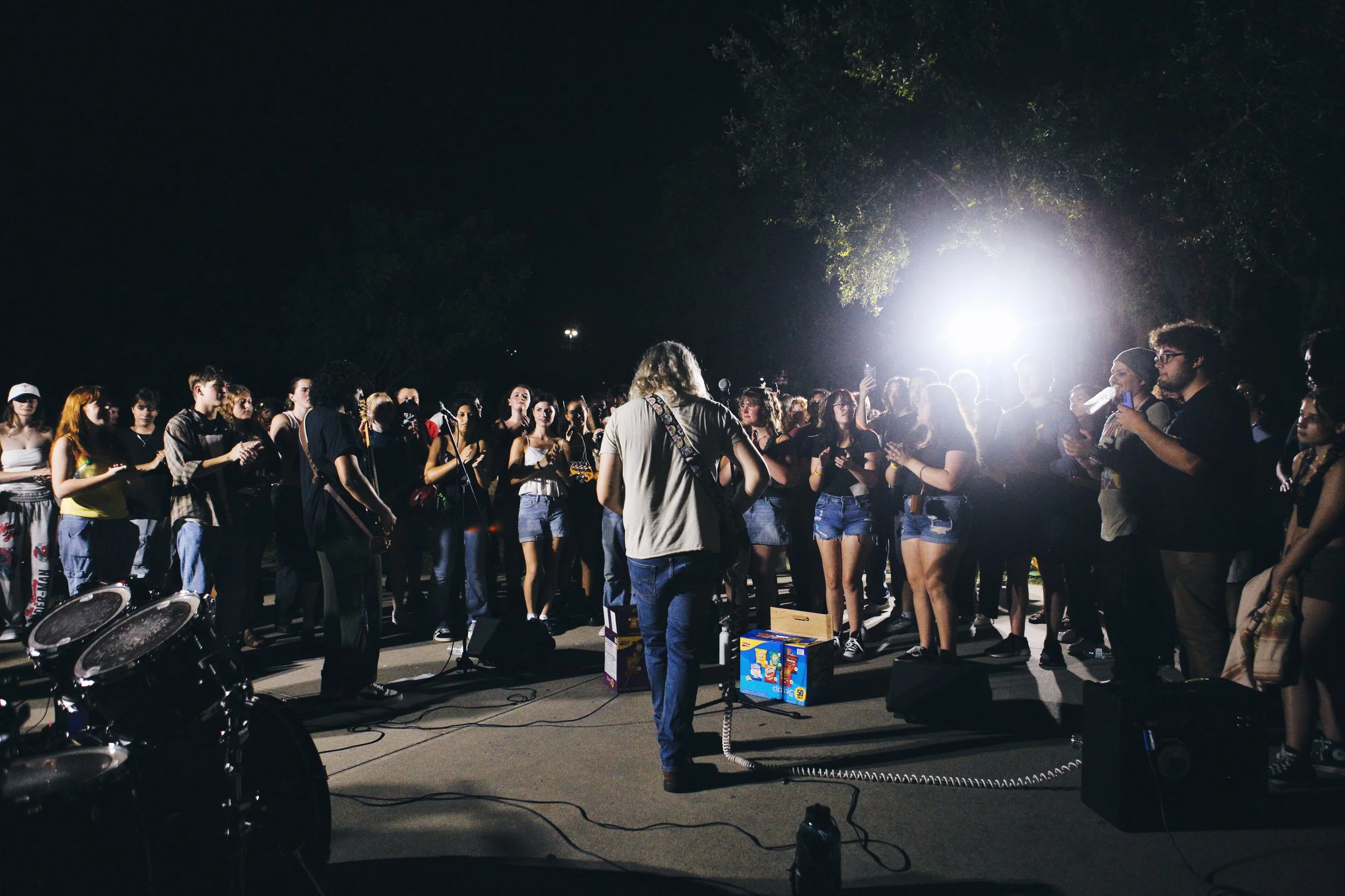 A musician performs with a band in front of a crowd at night, illuminated by bright lights. The audience is gathered closely, some holding drinks and recording the event with their phones.