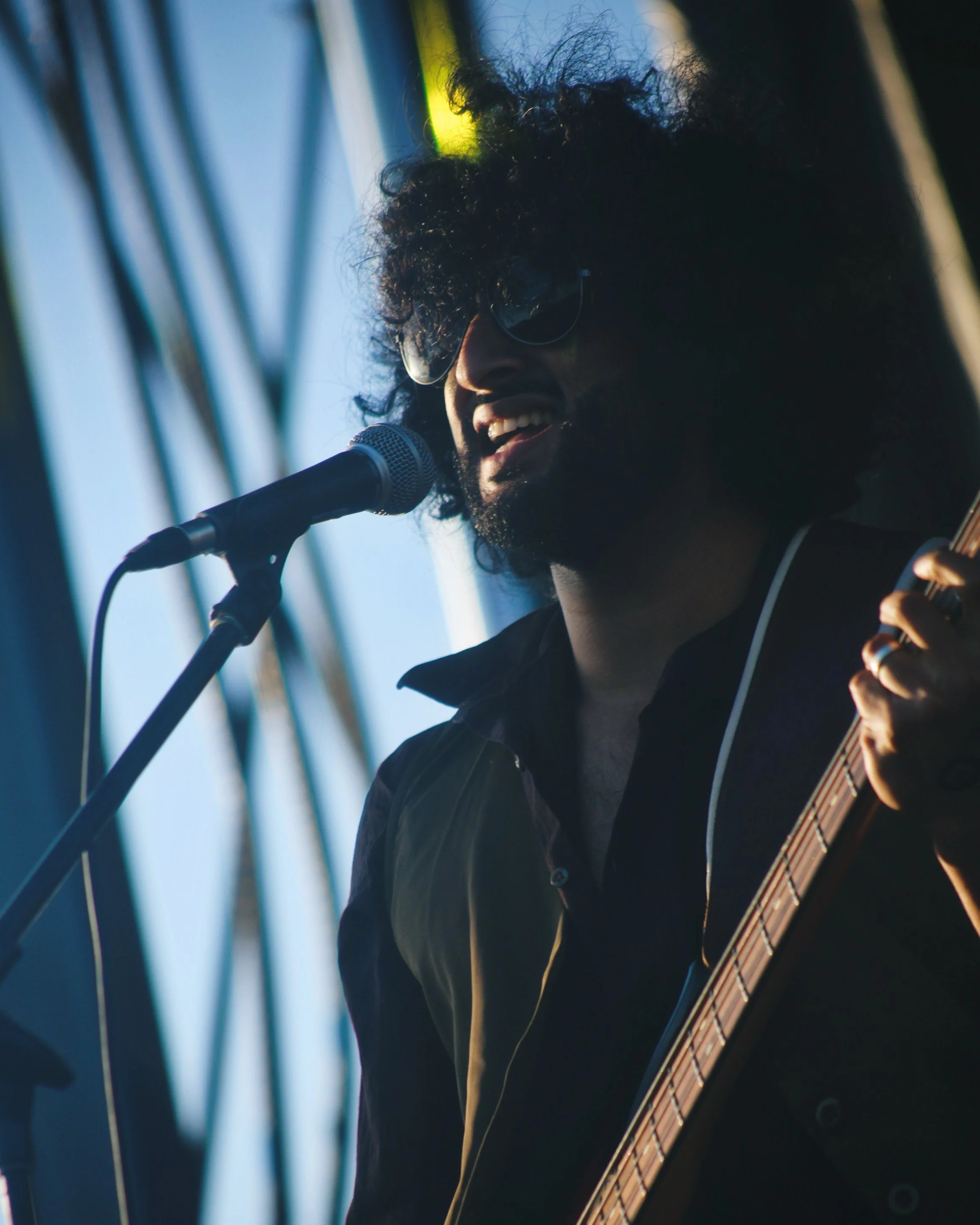 A man with curly hair and sunglasses singing into a microphone and playing guitar on stage.