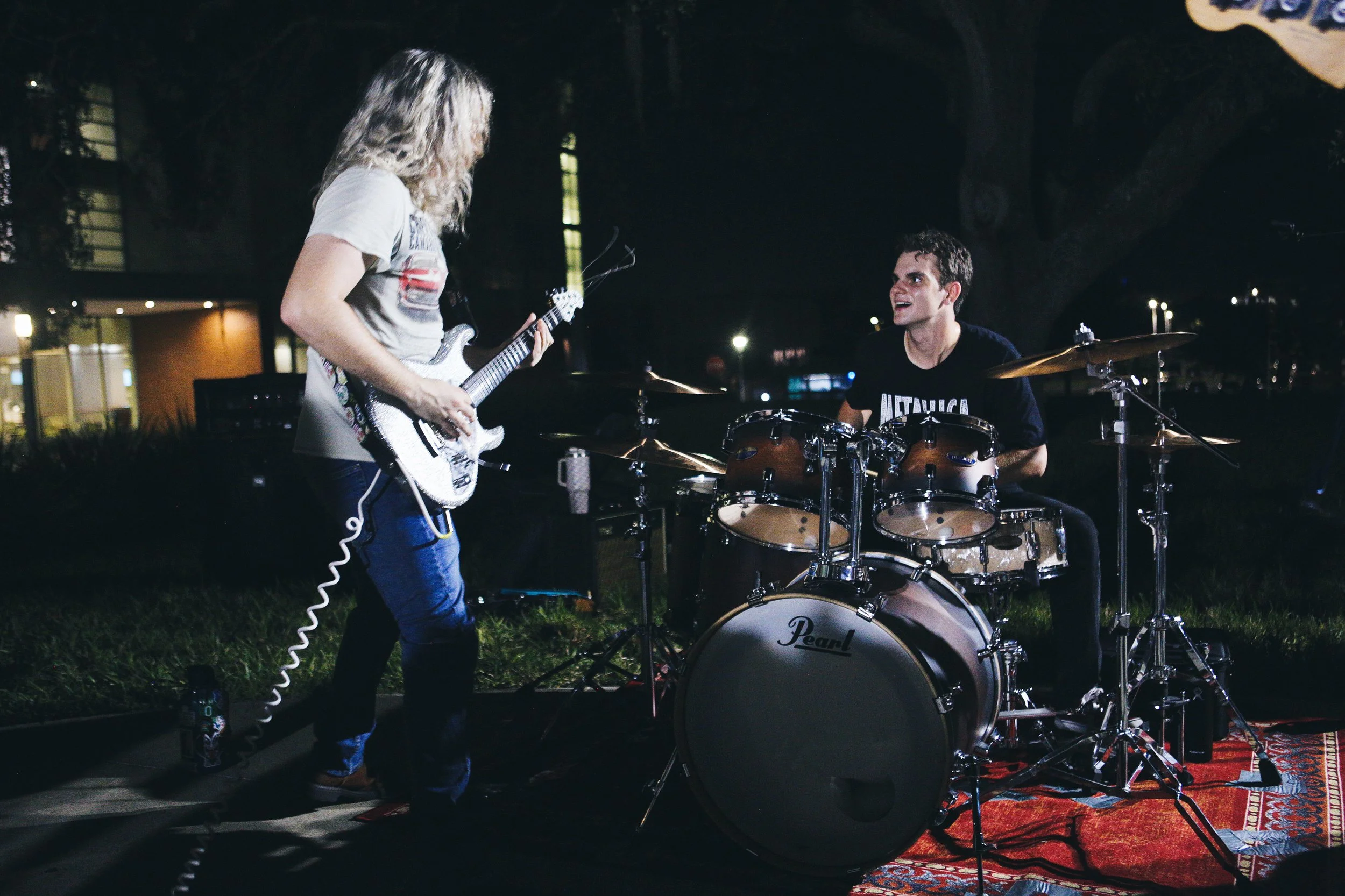 Two musicians performing outdoors at night, one playing an electric guitar and the other on a drum set, on a decorative rug.