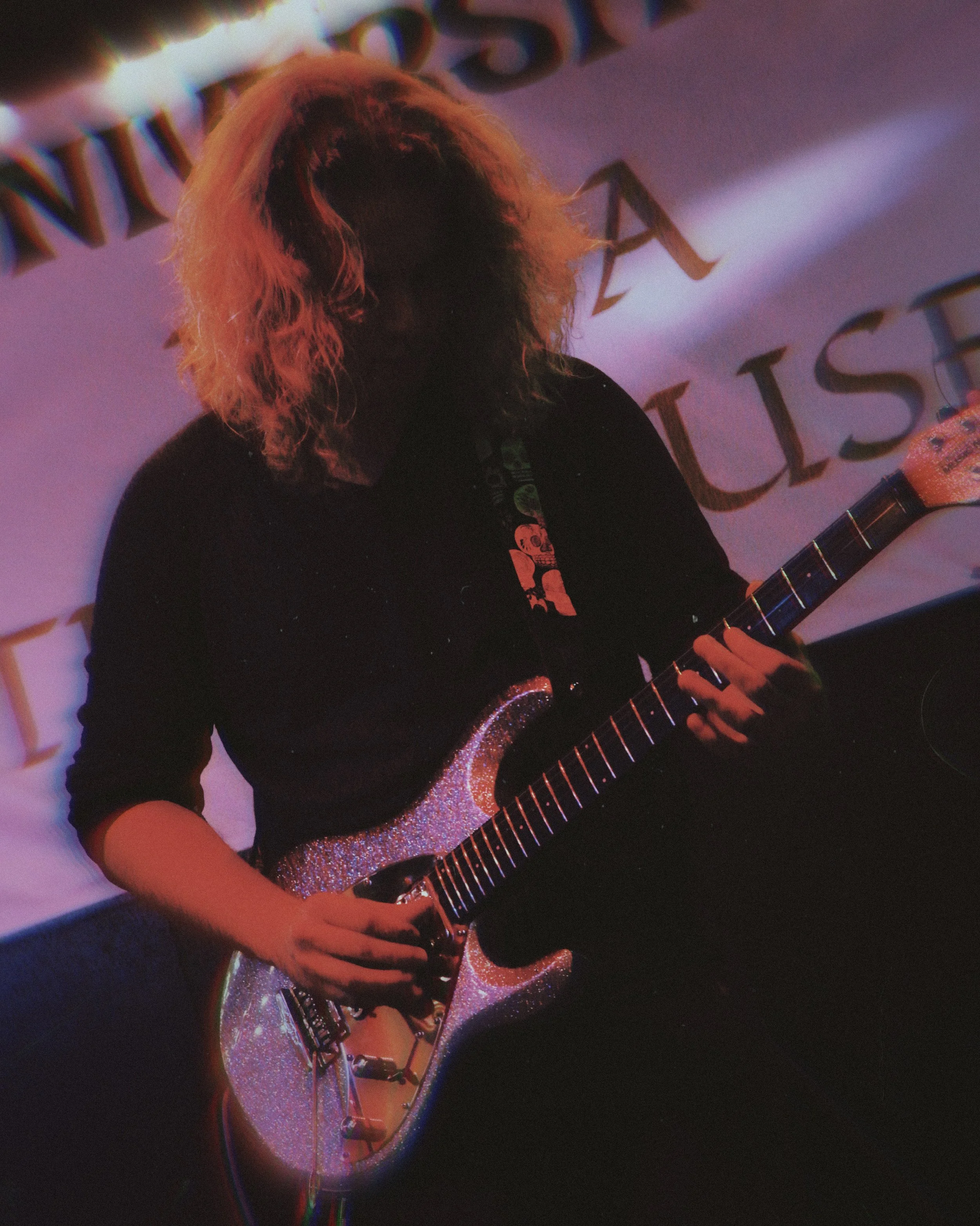 Person with long curly hair playing a glittery electric guitar on stage with a banner behind them.