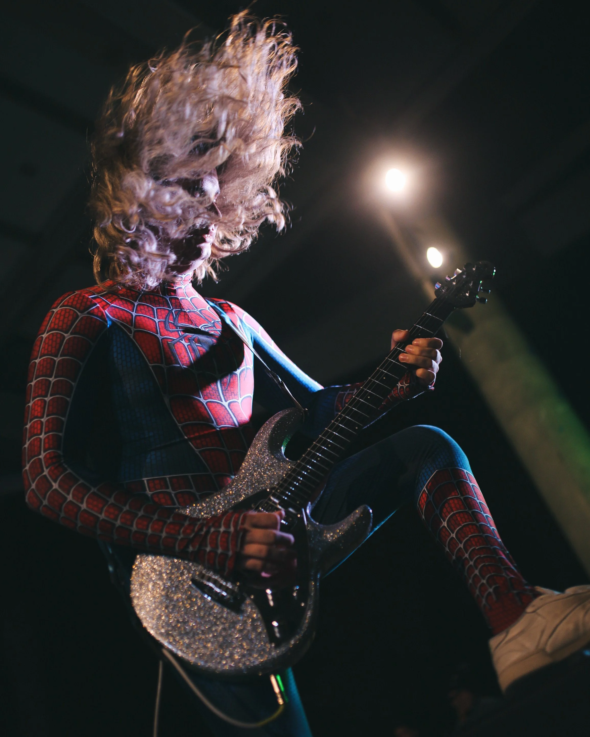 A woman dressed as Spider-Man playing an electric guitar on stage with lighting.