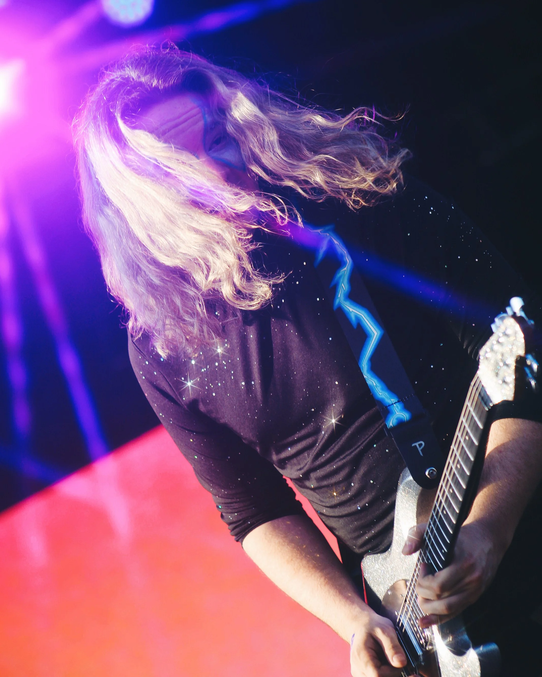 Person with long, wavy blonde hair playing an electric guitar on stage, illuminated by purple and pink stage lighting.